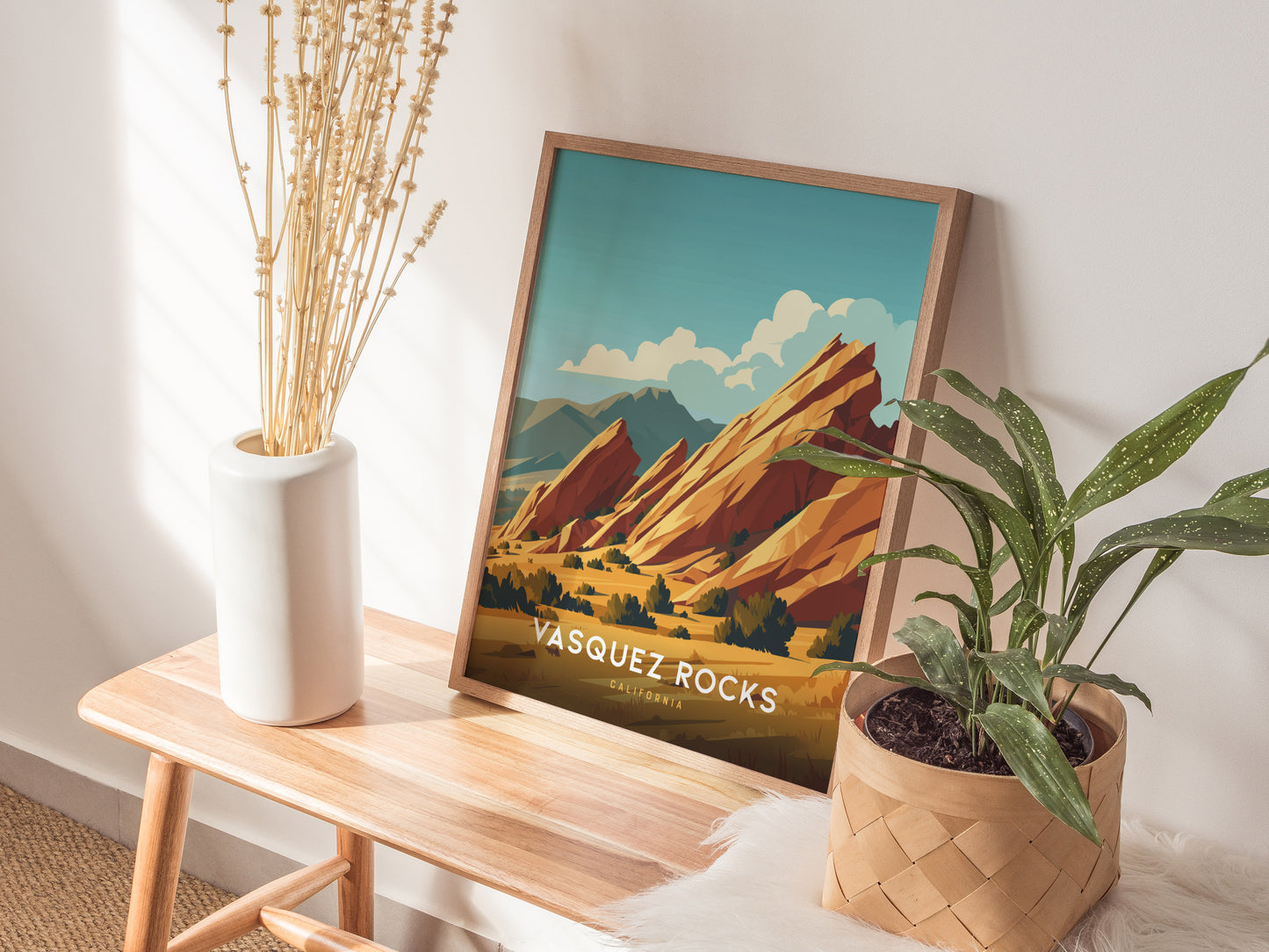 A framed Vasquez Rocks poster rests on a wooden bench beside a white vase with dried flowers and a potted green plant.