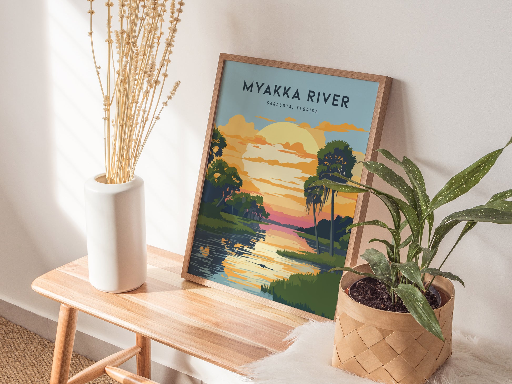 A Myakka River State Park poster with a sunset scene rests on a wooden bench, accompanied by a white vase with dried flowers and a potted plant.