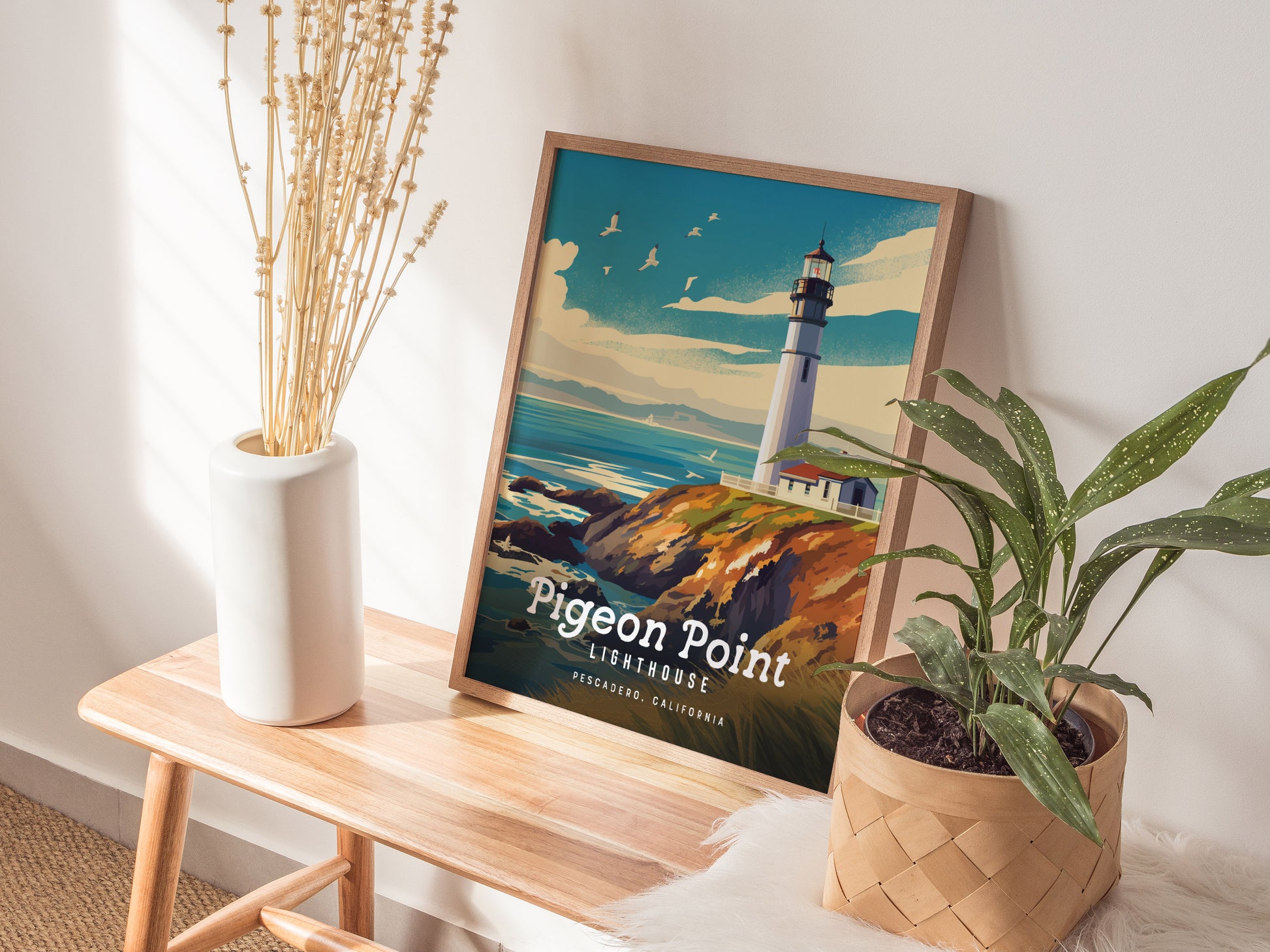 A framed poster of Pigeon Point Lighthouse rests on a wooden bench, surrounded by a white vase with dried flowers and a potted plant. Sunlight casts soft shadows.