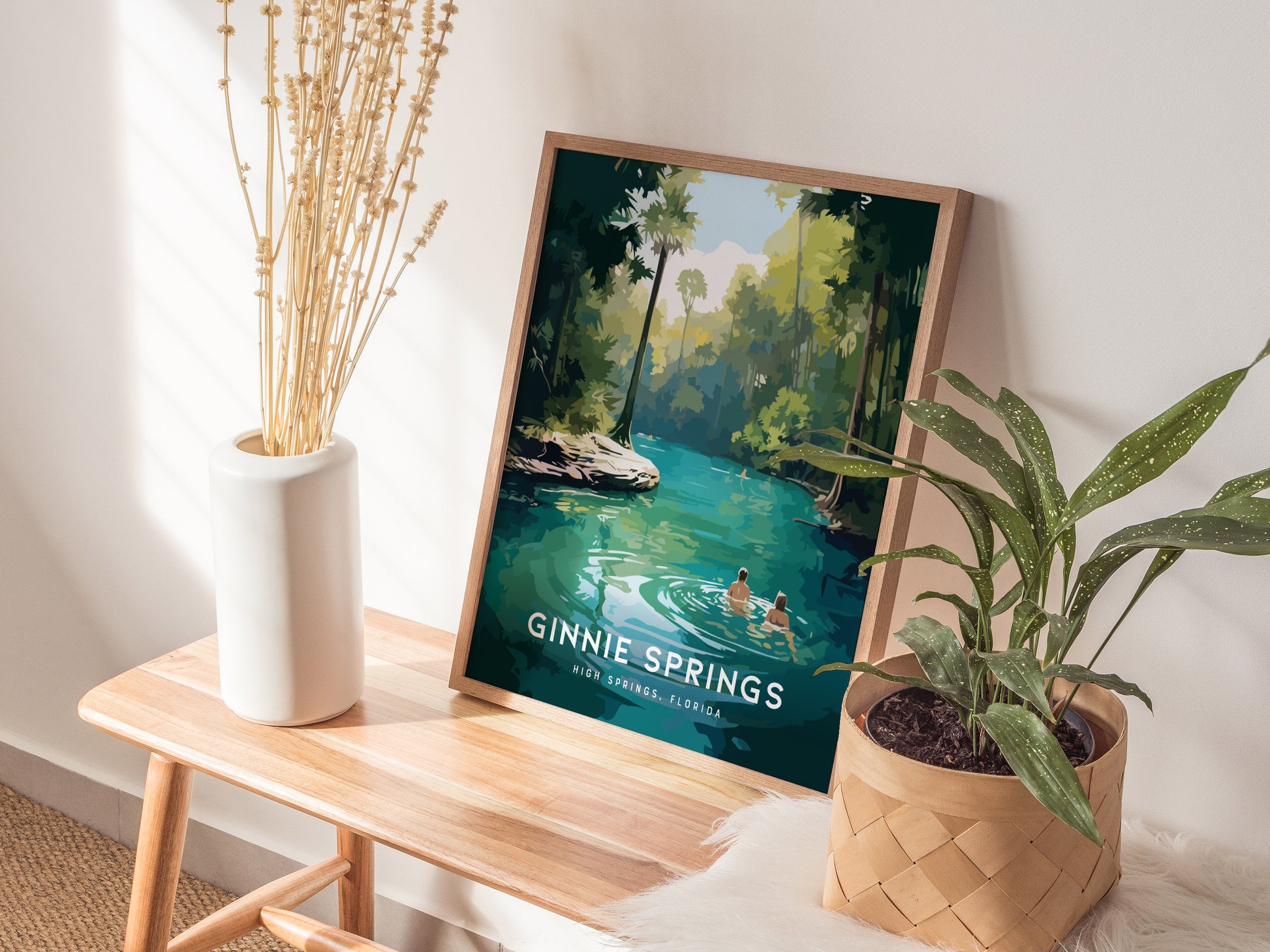 A framed Ginnie Springs poster, depicting a couple in turquoise waters surrounded by lush greenery, sits on a wooden table next to a white vase and a potted plant.