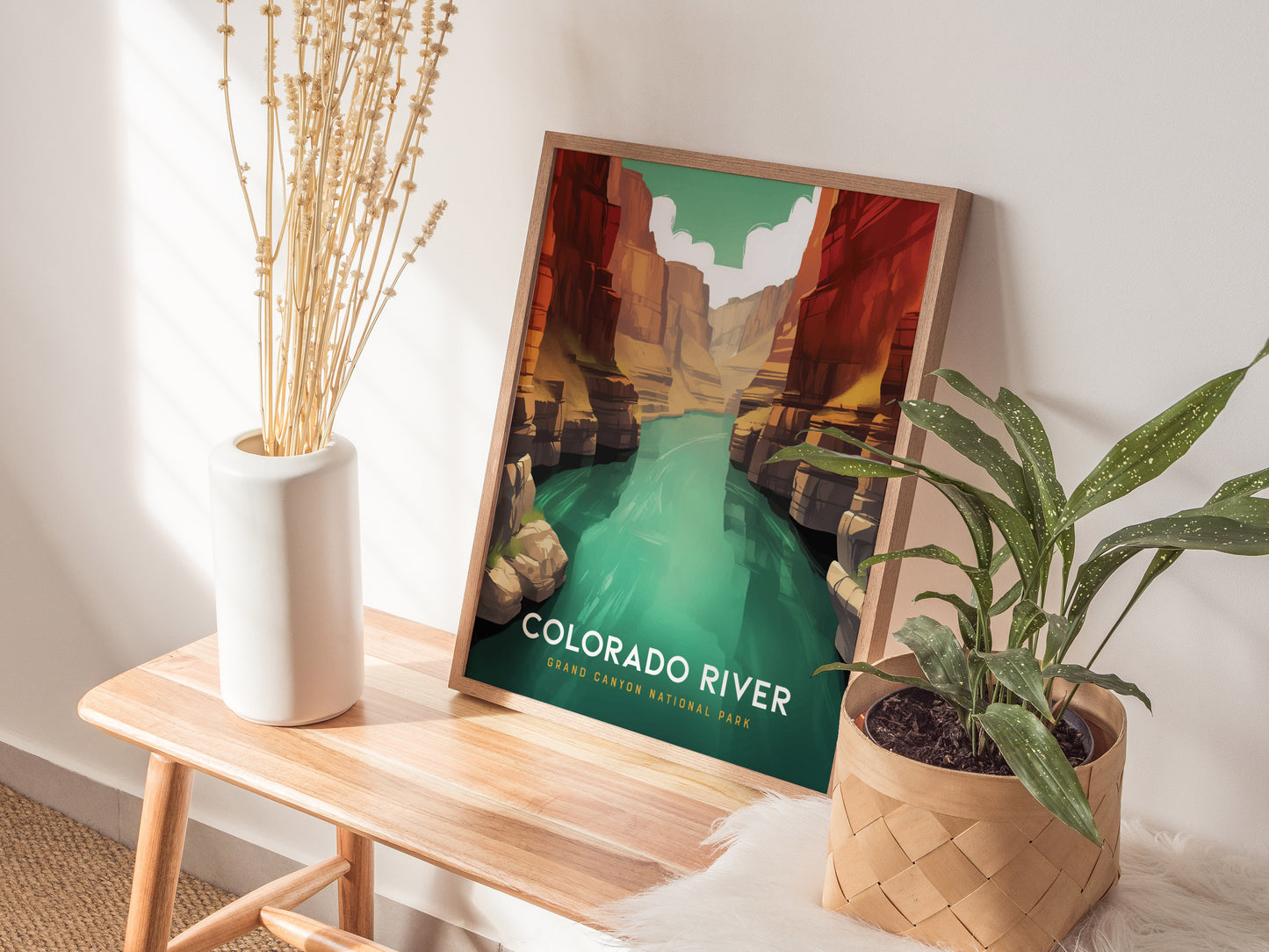 A framed poster of the Colorado River in the Grand Canyon is placed on a wooden bench, surrounded by a white vase with dried flowers and a potted plant.