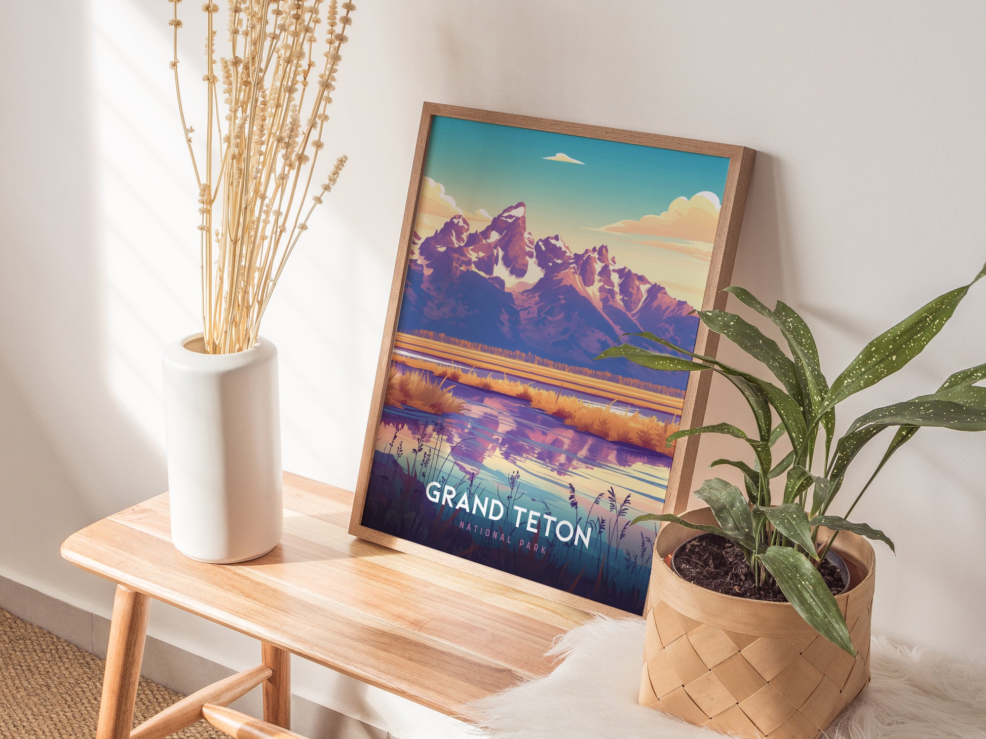 A framed Grand Teton National Park poster rests on a wooden bench, accompanied by a white vase with dried flowers and a potted plant. Sunlight casts shadows on the wall.