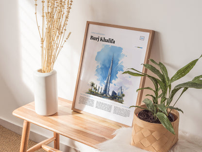 A framed poster of the Burj Khalifa rests on a wooden bench, accompanied by a white vase with dried branches and a potted green plant. Natural light illuminates the scene.