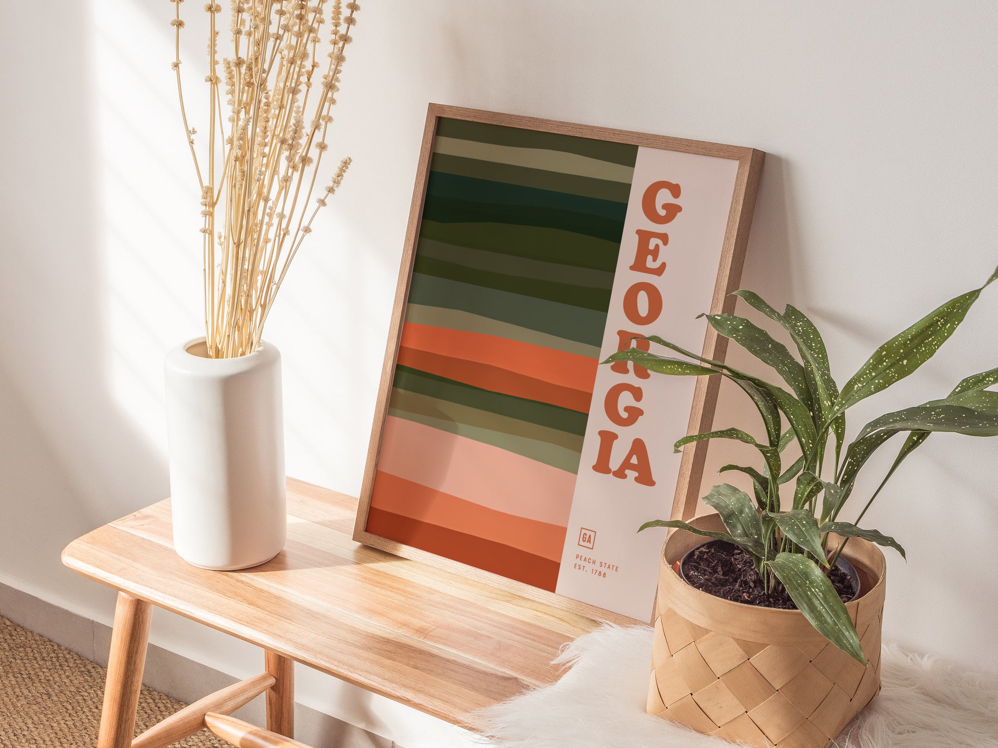 A Georgia-themed poster with bold typography and earthy horizontal lines rests on a wooden bench alongside a white vase with dried flowers and a potted green plant.