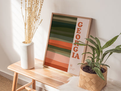 A Georgia-themed poster with bold typography and earthy horizontal lines rests on a wooden bench alongside a white vase with dried flowers and a potted green plant.