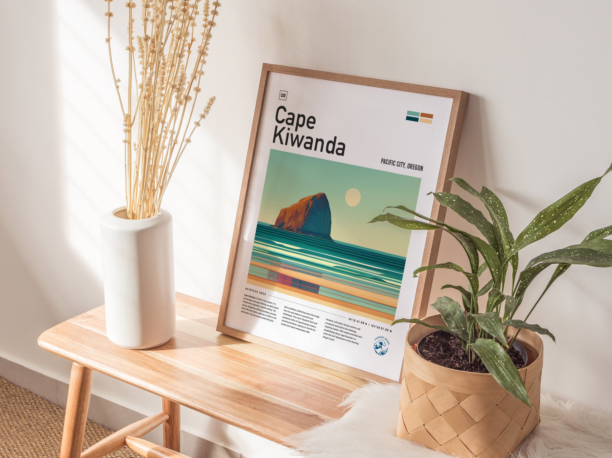 A framed Cape Kiwanda poster rests on a wooden bench, accompanied by a white vase with dried flowers and a potted plant.