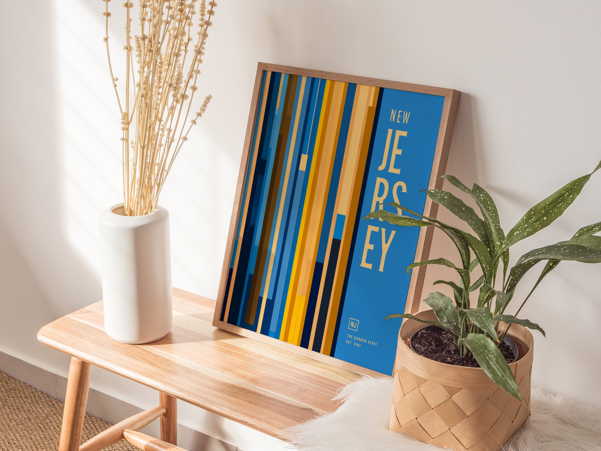 A New Jersey poster with bold vertical lines and text is displayed on a wooden bench, accompanied by a white vase with dried stems and a potted plant.