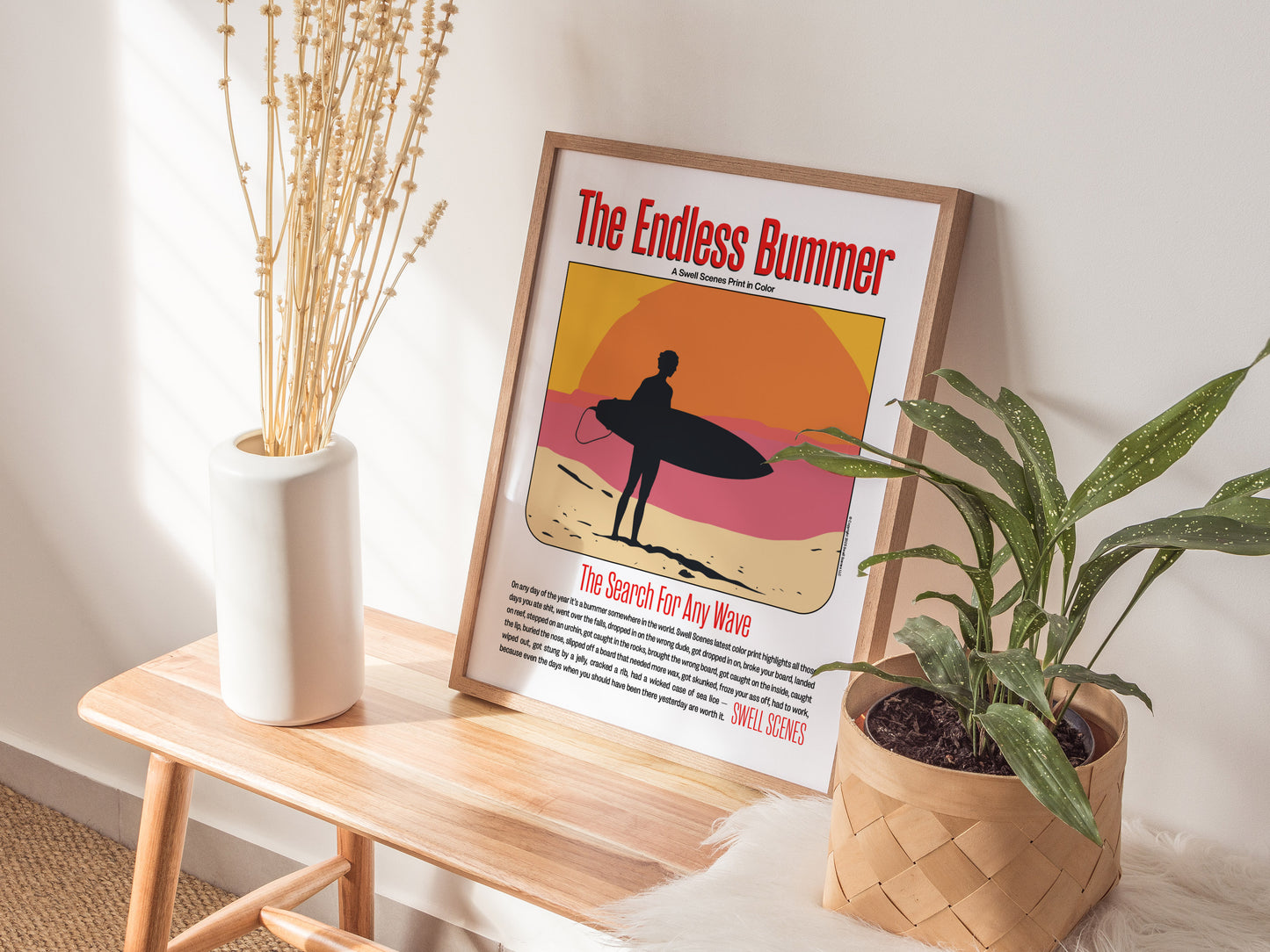 A framed The Endless Bummer poster rests on a wooden bench, accompanied by a plant in a woven basket and a white vase with tall dried flowers.