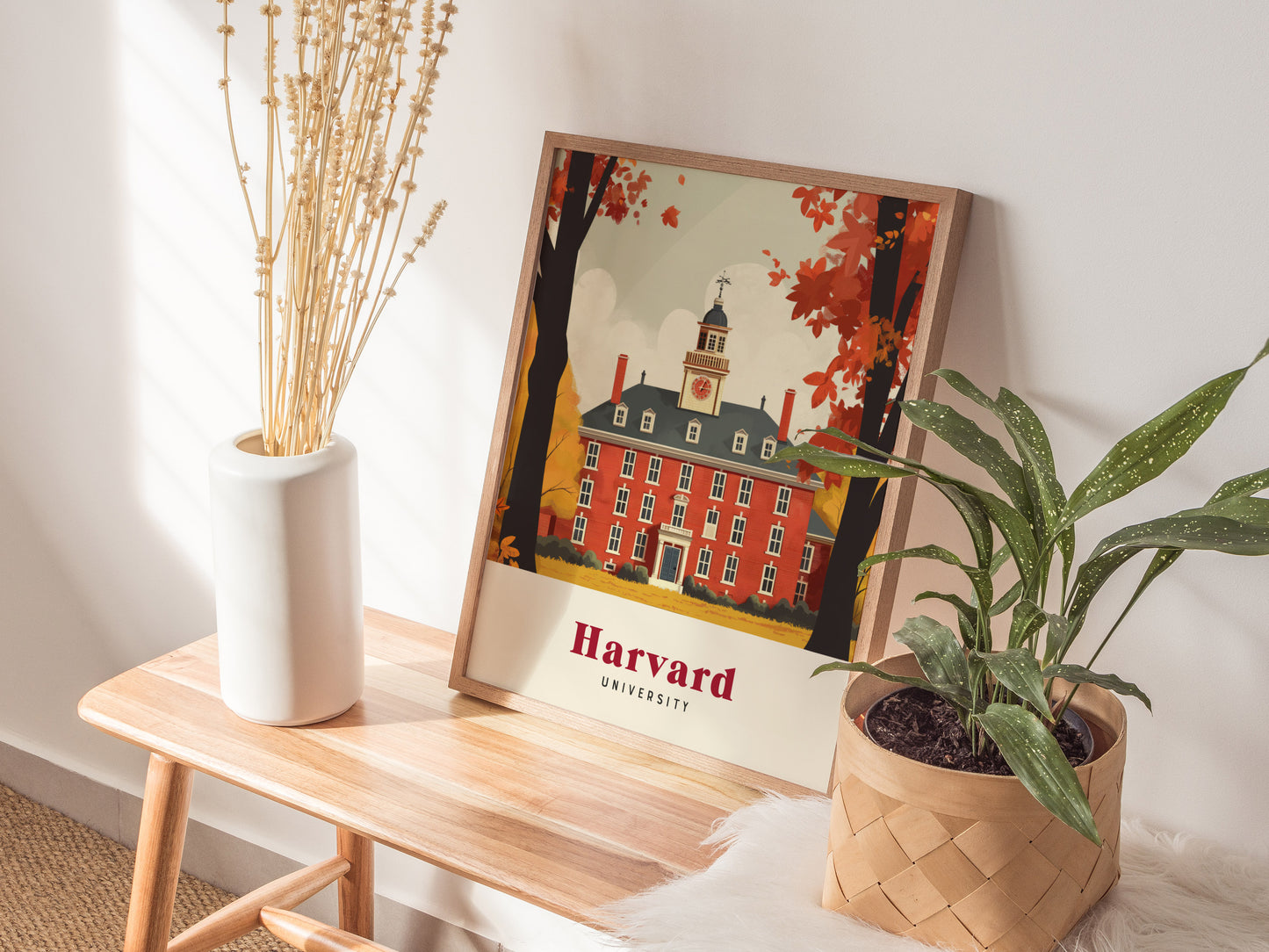 A framed Harvard University poster rests on a wooden bench, surrounded by a white vase with dried flowers and a potted green plant, creating a cozy decor scene.
