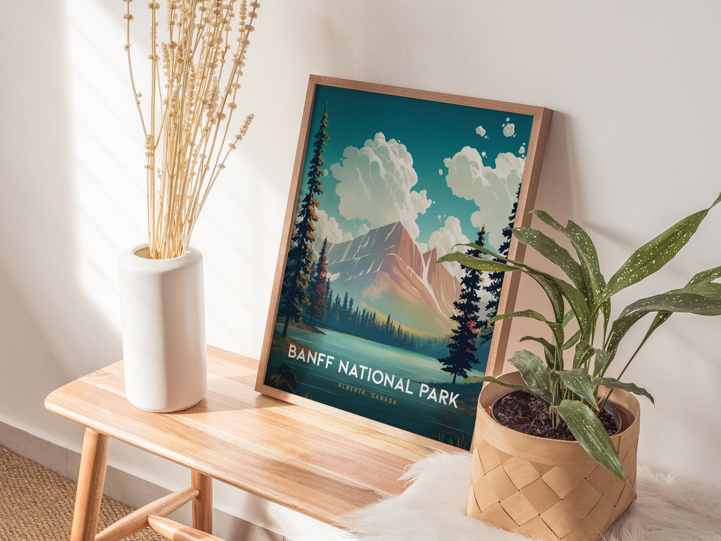 A framed Banff National Park poster rests on a wooden bench, adorned with a potted plant and a vase of dried flowers, creating a cozy, nature-inspired setting.