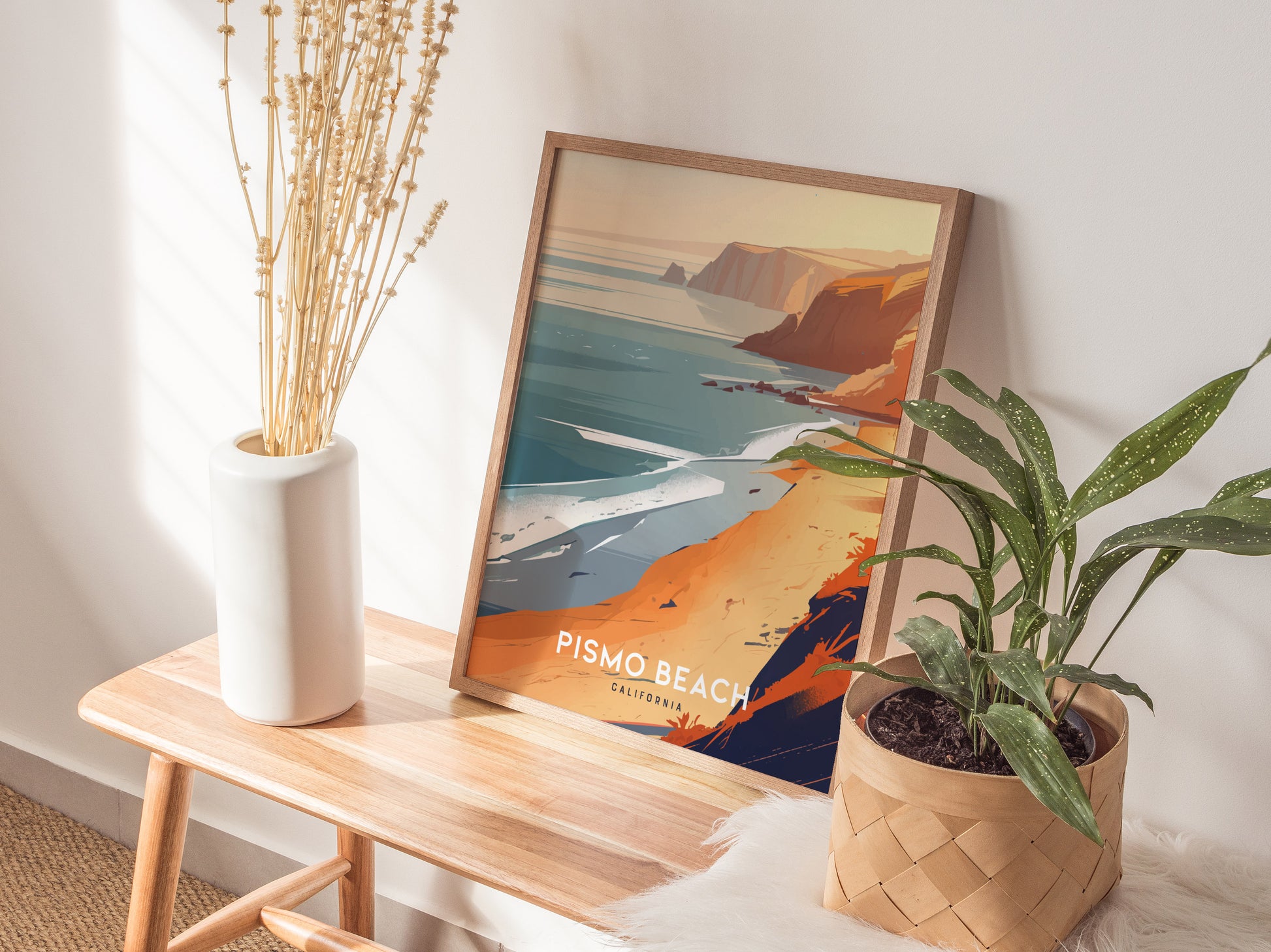 A framed Pismo Beach poster depicting a colorful coastal scene sits on a wooden bench. Nearby, a white vase with dried flowers and a potted plant add decorative elements.