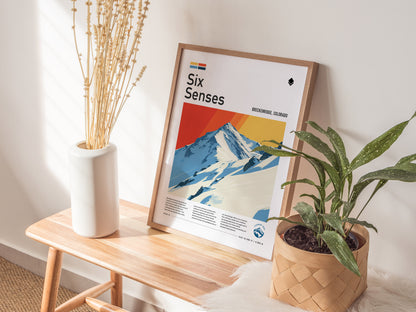 A framed poster of Breckenridges Peak 6 leans against a wall, featuring vibrant mountain artwork. It sits on a wooden bench beside a vase of dried flowers and a potted.