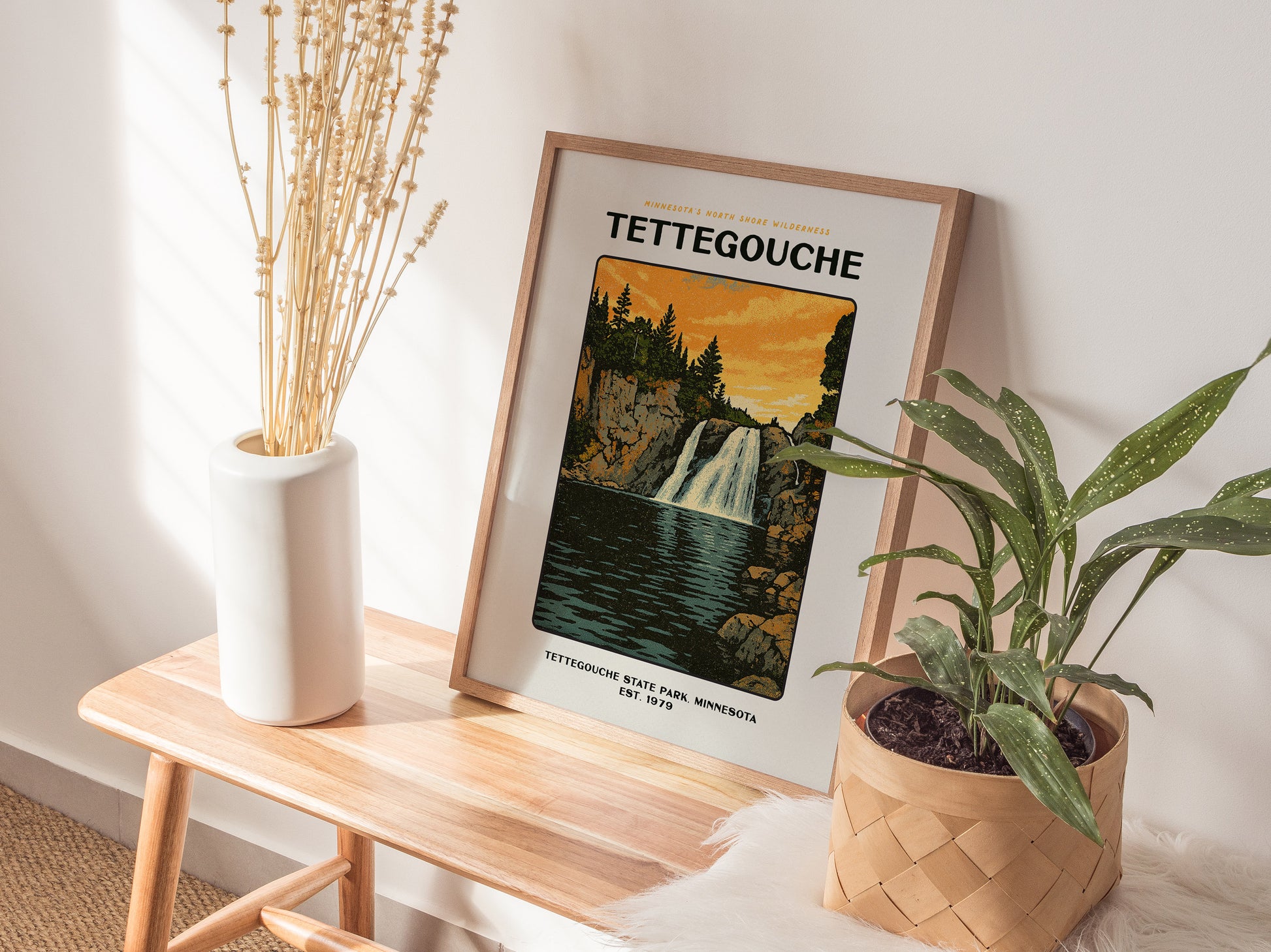 A framed Tettegouche poster featuring a waterfall is displayed on a wooden bench, next to a white vase with dried flowers and a green plant in a woven basket.