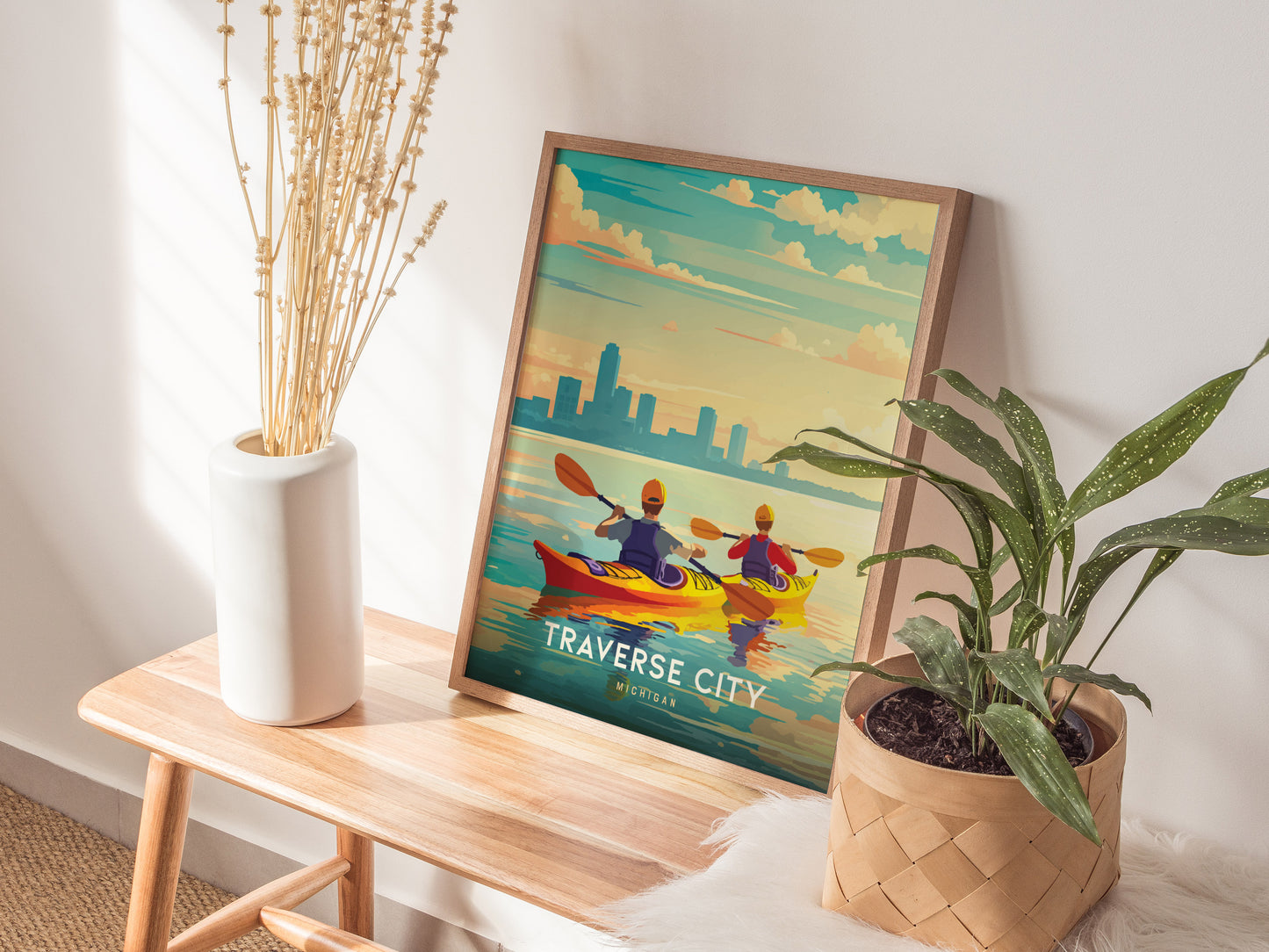 A framed Traverse City poster is displayed on a wooden bench, featuring kayakers on Grand Traverse Bay. Its surrounded by a plant in a woven basket and a vase with.