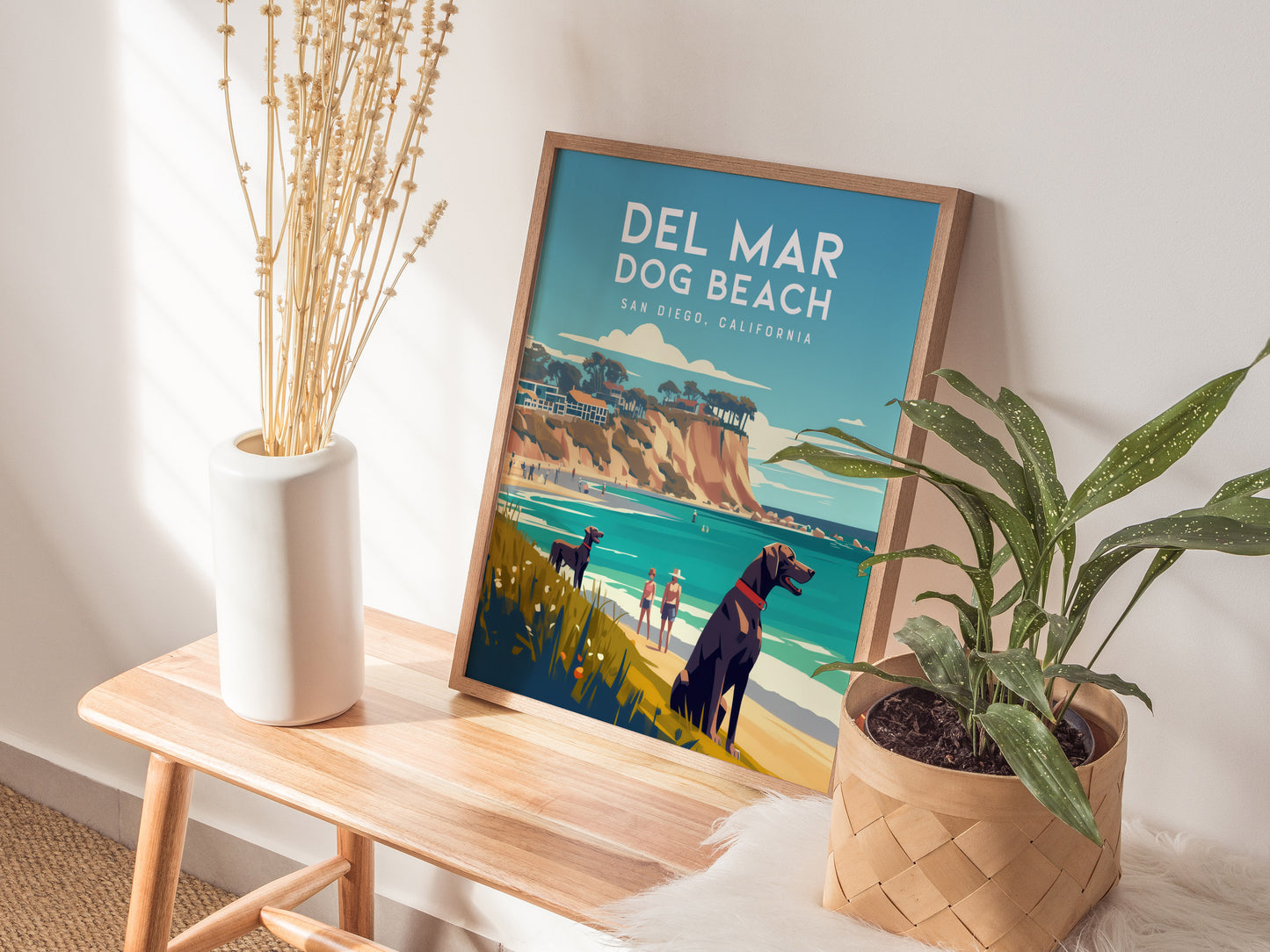 A framed Del Mar Dog Beach poster sits on a wooden bench, surrounded by a white vase with dried flowers and a green potted plant, evoking a coastal vibe.