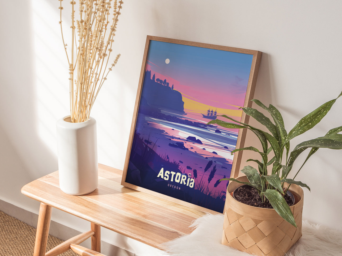 A framed poster depicting a colorful sunset over a coastal landscape with a pirate ship, titled Astoria, is placed on a wooden bench beside a plant and vase.