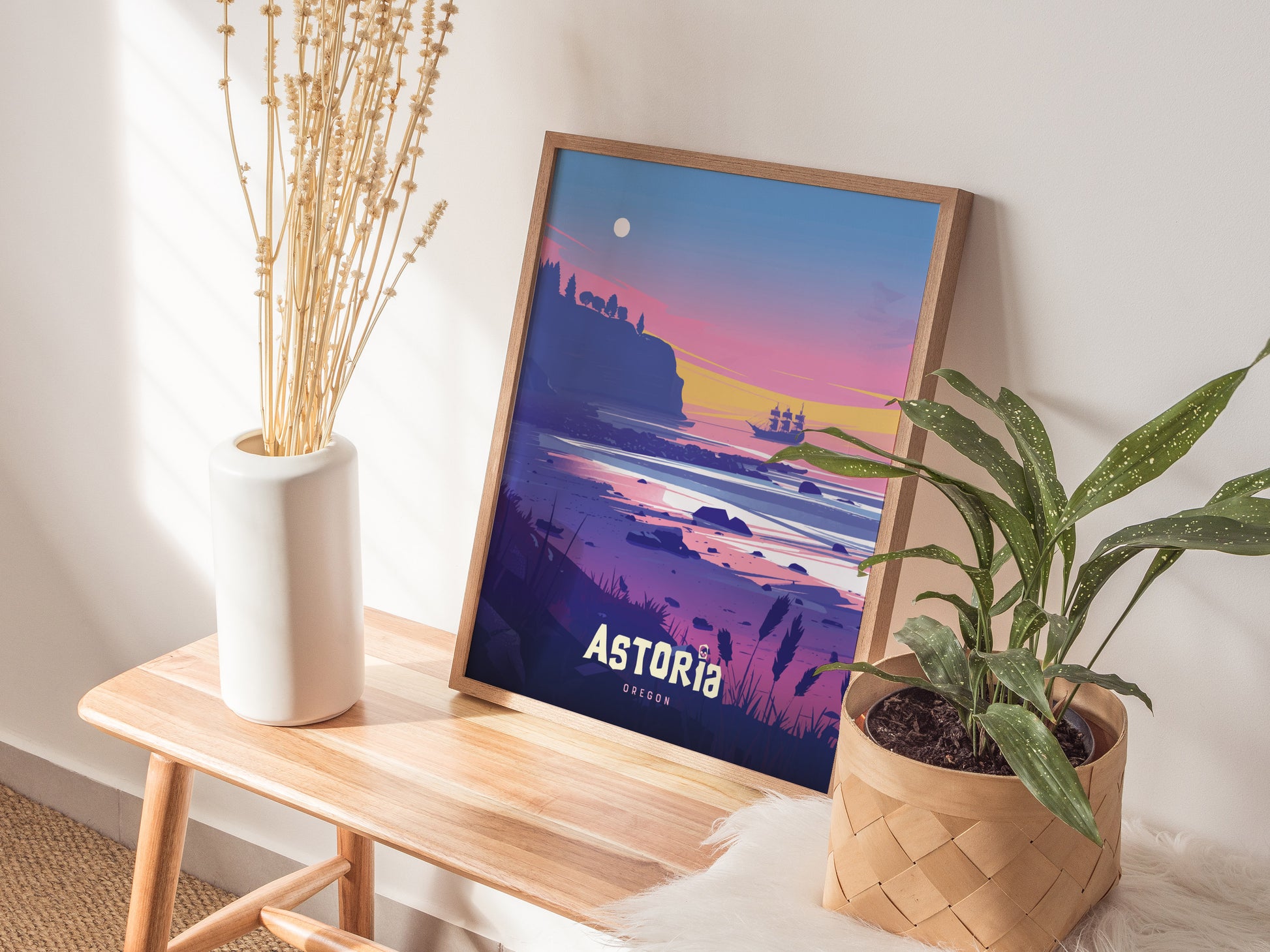 A framed poster depicting a colorful sunset over a coastal landscape with a pirate ship, titled Astoria, is placed on a wooden bench beside a plant and vase.