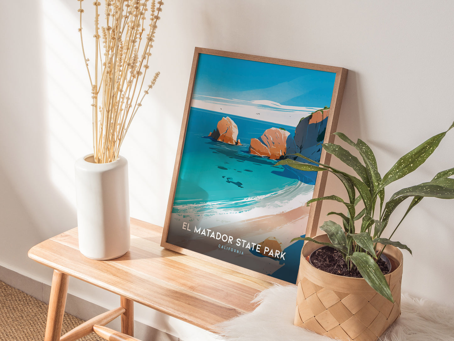 A framed El Matador State Park poster leans against a wall on a wooden bench, accompanied by a white vase with dried flowers and a potted plant.
