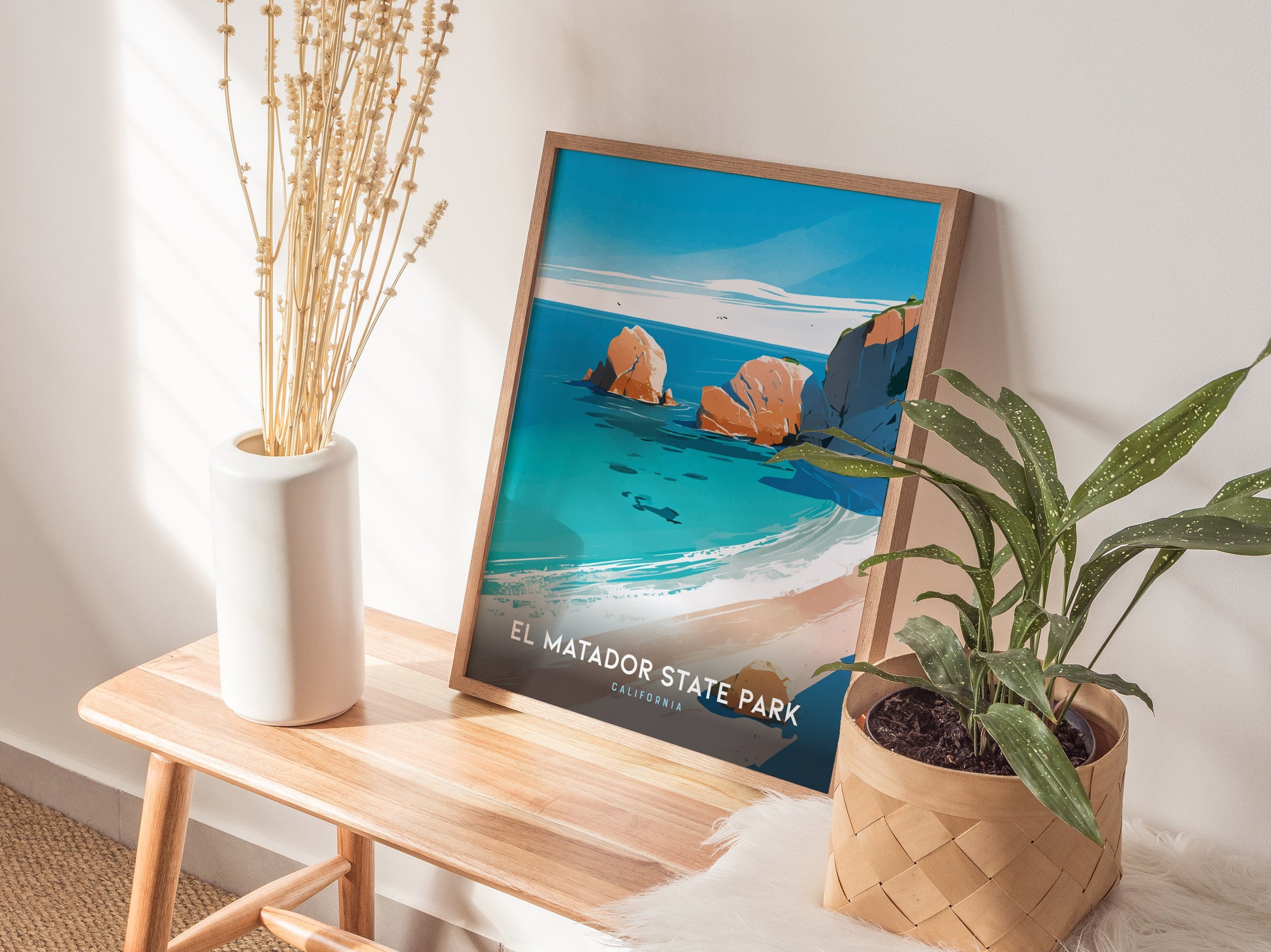 A framed El Matador State Park poster leans against a wall on a wooden bench, accompanied by a white vase with dried flowers and a potted plant.
