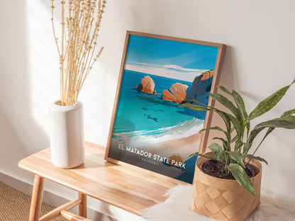 A framed El Matador State Park poster leans against a wall on a wooden bench, accompanied by a white vase with dried flowers and a potted plant.
