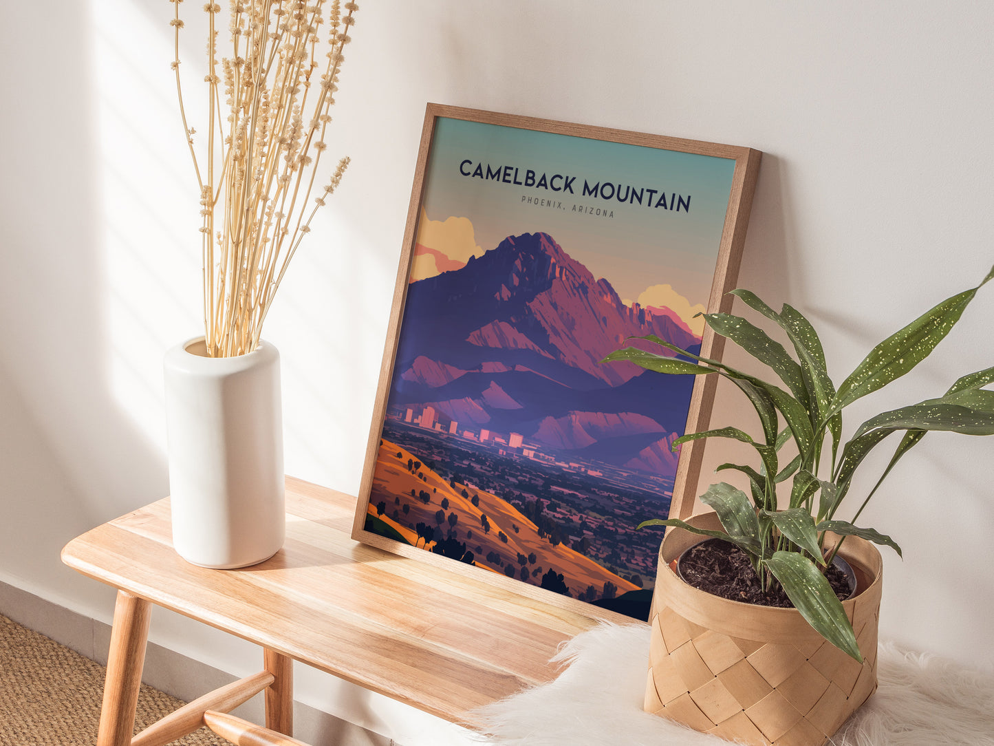 A framed Camelback Mountain poster sits on a wooden bench, accompanied by a white vase with dried plants and a potted green plant, creating a cozy decor setting.