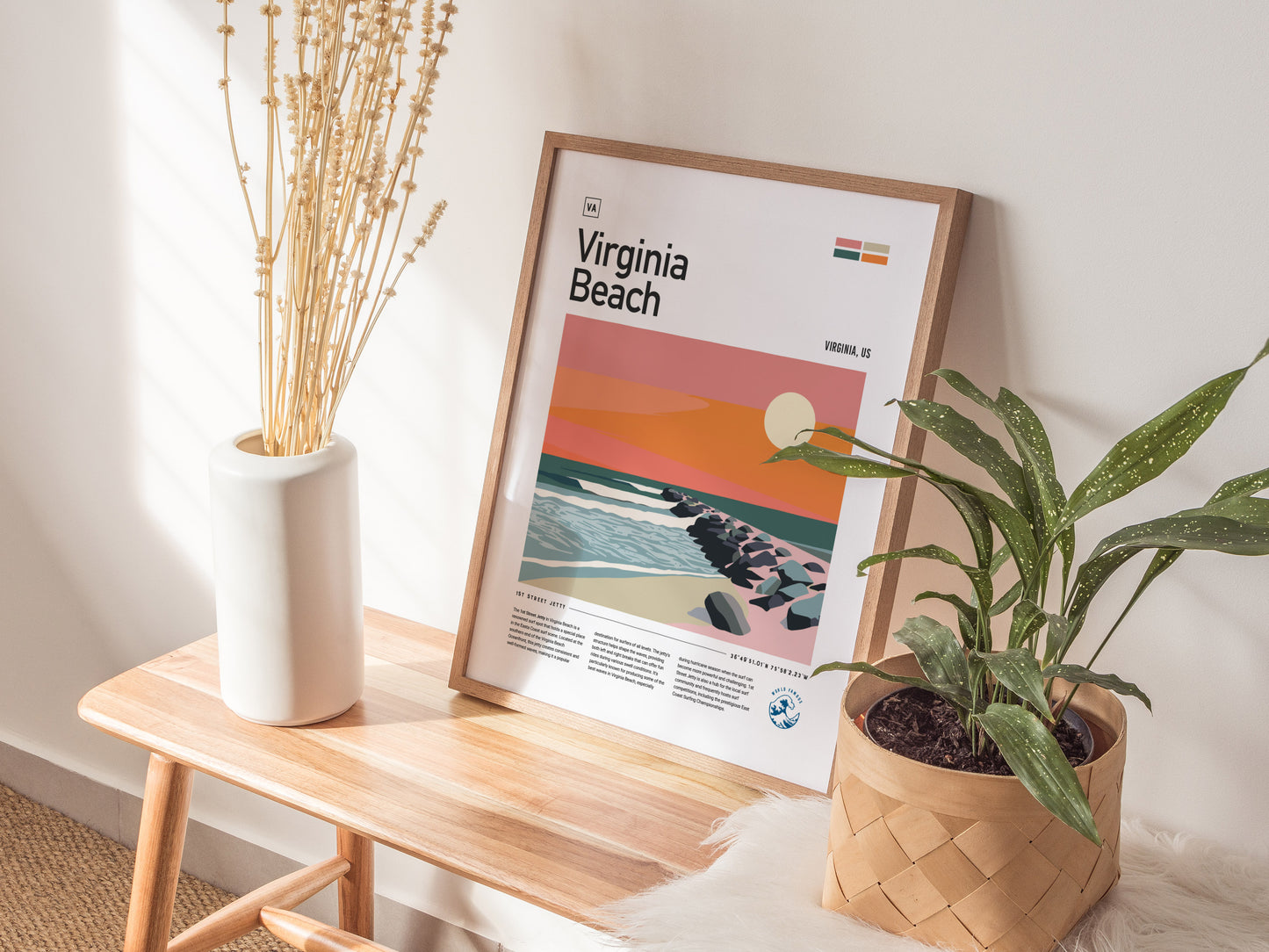 A framed Virginia Beach poster featuring a colorful sunset and ocean scene is displayed on a wooden bench next to a vase of dried flowers and a potted plant.