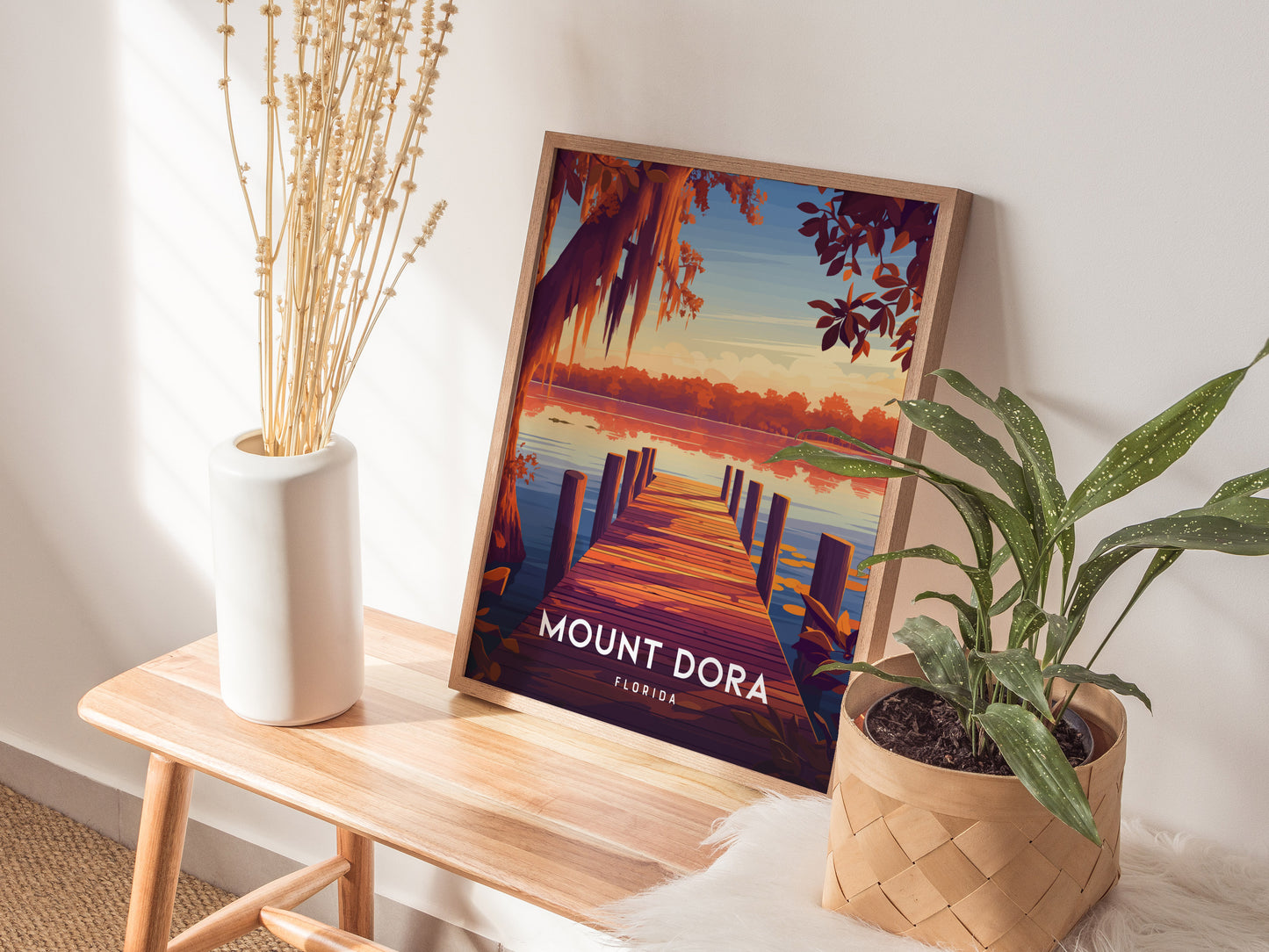 A framed Mount Dora, Florida poster rests on a wooden bench, surrounded by a white vase with dried stems and a potted plant. Sunlight casts shadows on the wall.