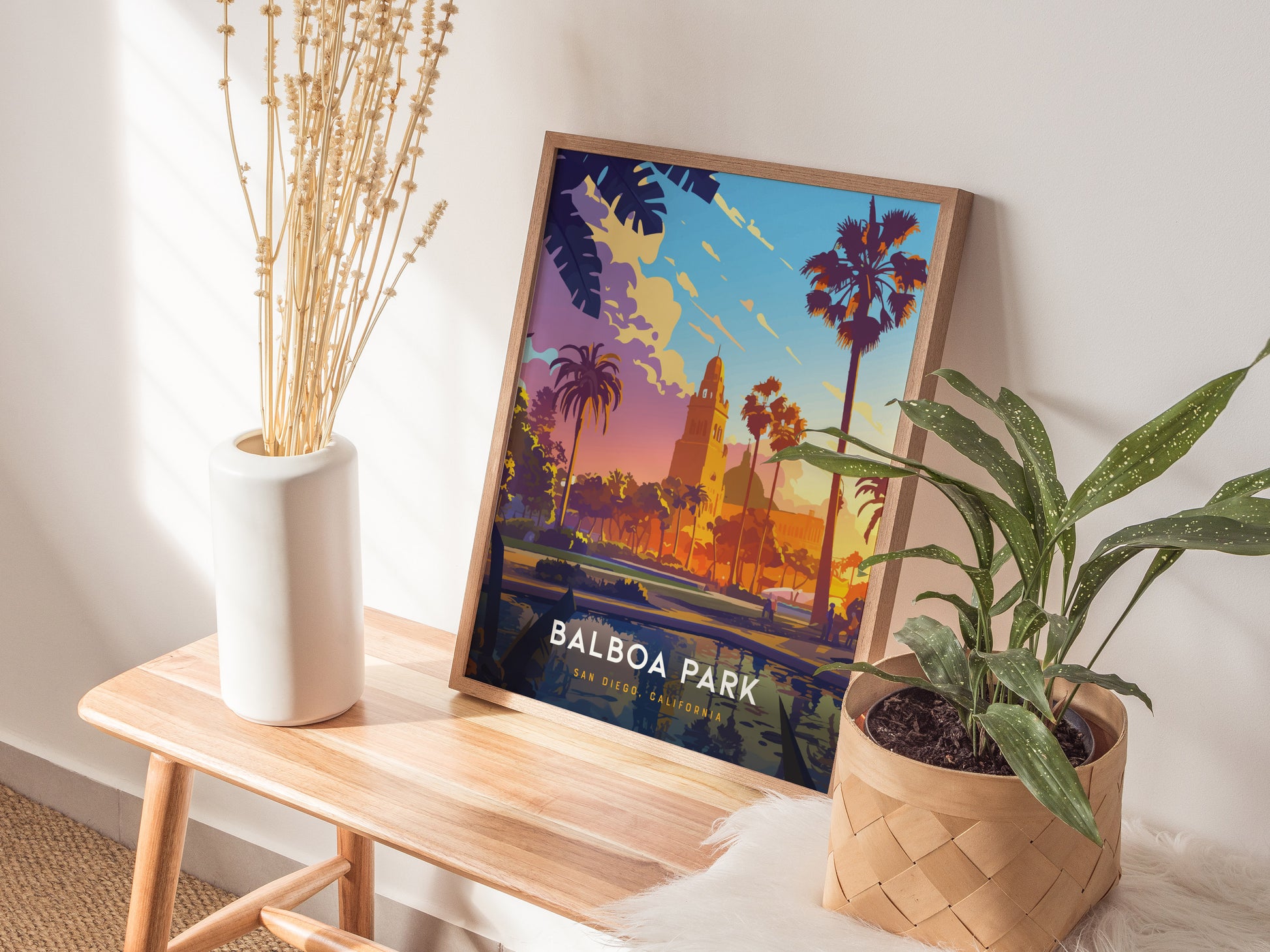 A colorful Balboa Park poster rests on a wooden table, accompanied by a white vase with dried stems and a potted green plant.