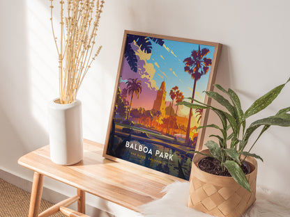 A colorful Balboa Park poster rests on a wooden table, accompanied by a white vase with dried stems and a potted green plant.