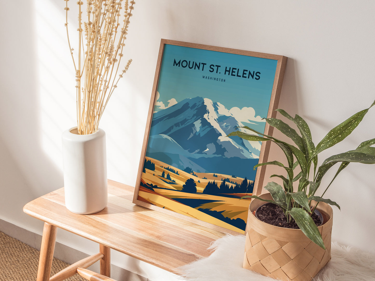 A framed Mount St. Helens poster rests on a wooden bench beside a white vase with dried stems and a potted plant. Sunlight casts soft shadows on the wall.