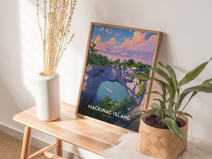 A framed poster of Mackinac Islands Arch Rock sits on a wooden bench, surrounded by a white vase with dried flowers and a leafy plant in a woven basket.