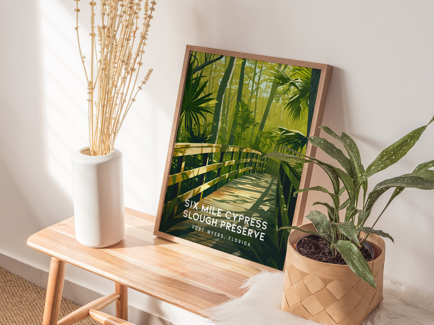 A framed poster of Six Mile Cypress Slough Preserve rests on a wooden bench, surrounded by a white vase with dried flowers and a potted plant.