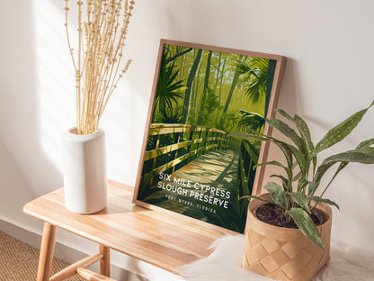 A framed poster of Six Mile Cypress Slough Preserve rests on a wooden bench, surrounded by a white vase with dried flowers and a potted plant.