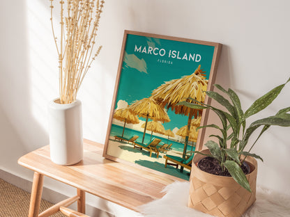 A framed poster of Marco Island, Florida, featuring beach lounge chairs and umbrellas, sits on a wooden table beside a vase with dried flowers and a potted plant.