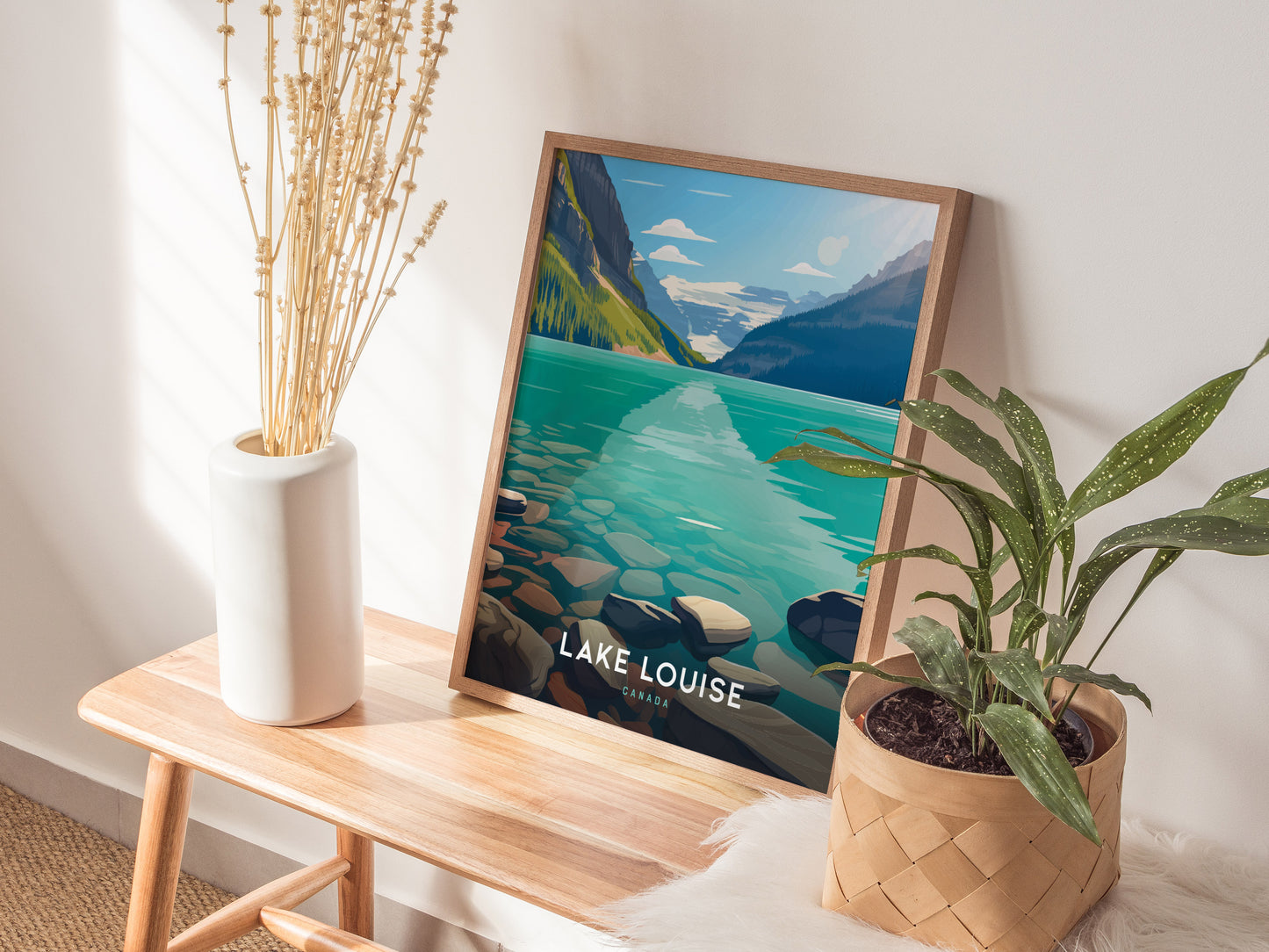 A framed Lake Louise poster rests on a wooden bench beside a white vase with dried flowers and a potted green plant, creating a serene, natural decor setting.