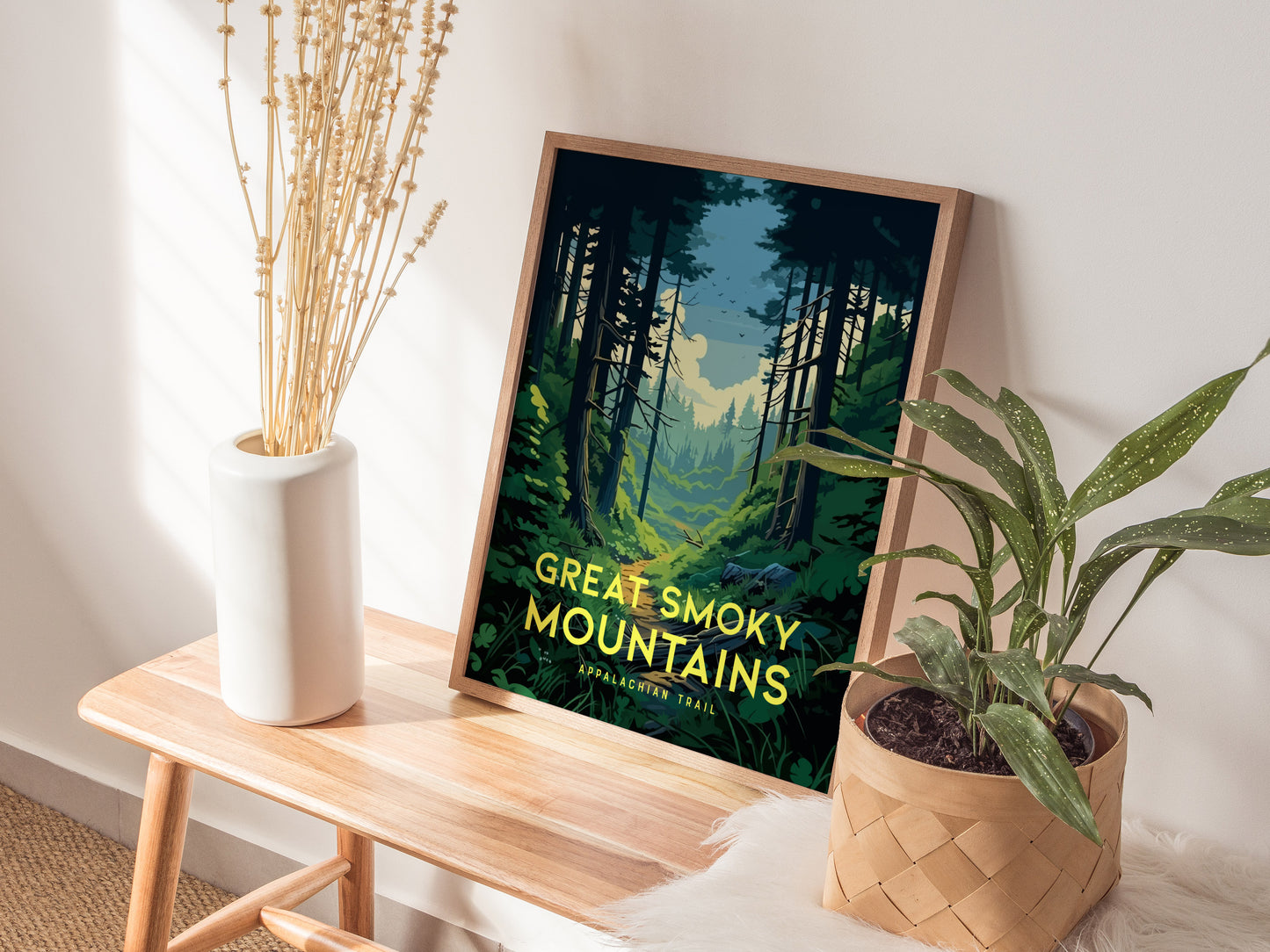 A framed Great Smoky Mountains poster rests on a wooden bench, accompanied by a white vase with dried flowers and a potted plant, creating a serene display.
