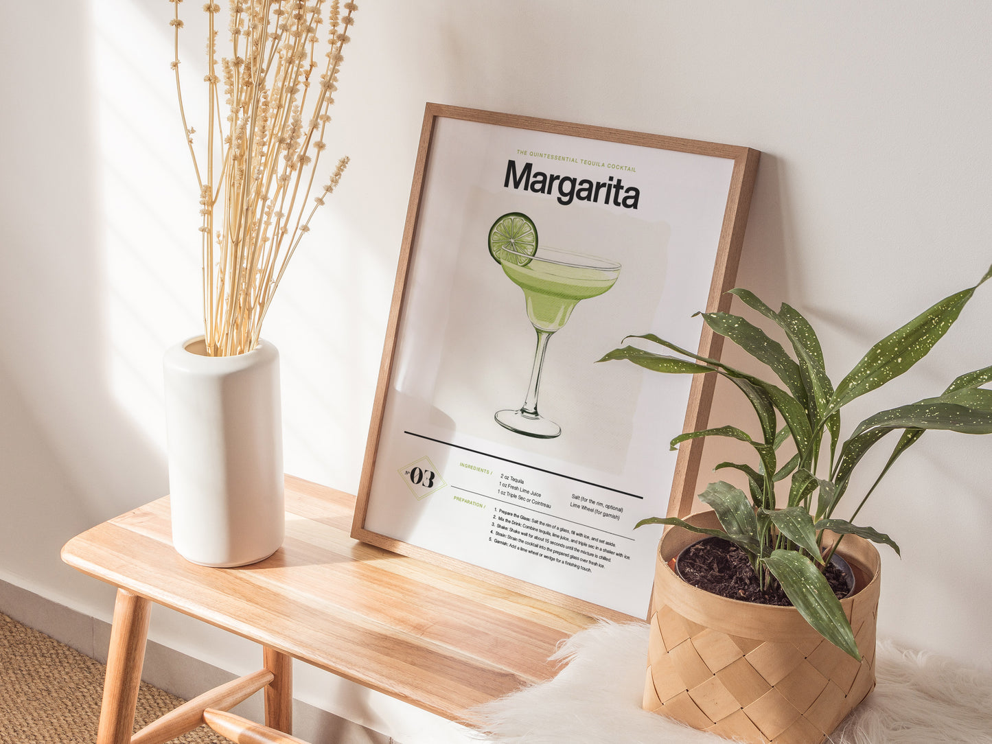A framed margarita cocktail poster rests on a wooden bench, flanked by a white vase with dried stems and a potted green plant. Sunlight casts subtle shadows.