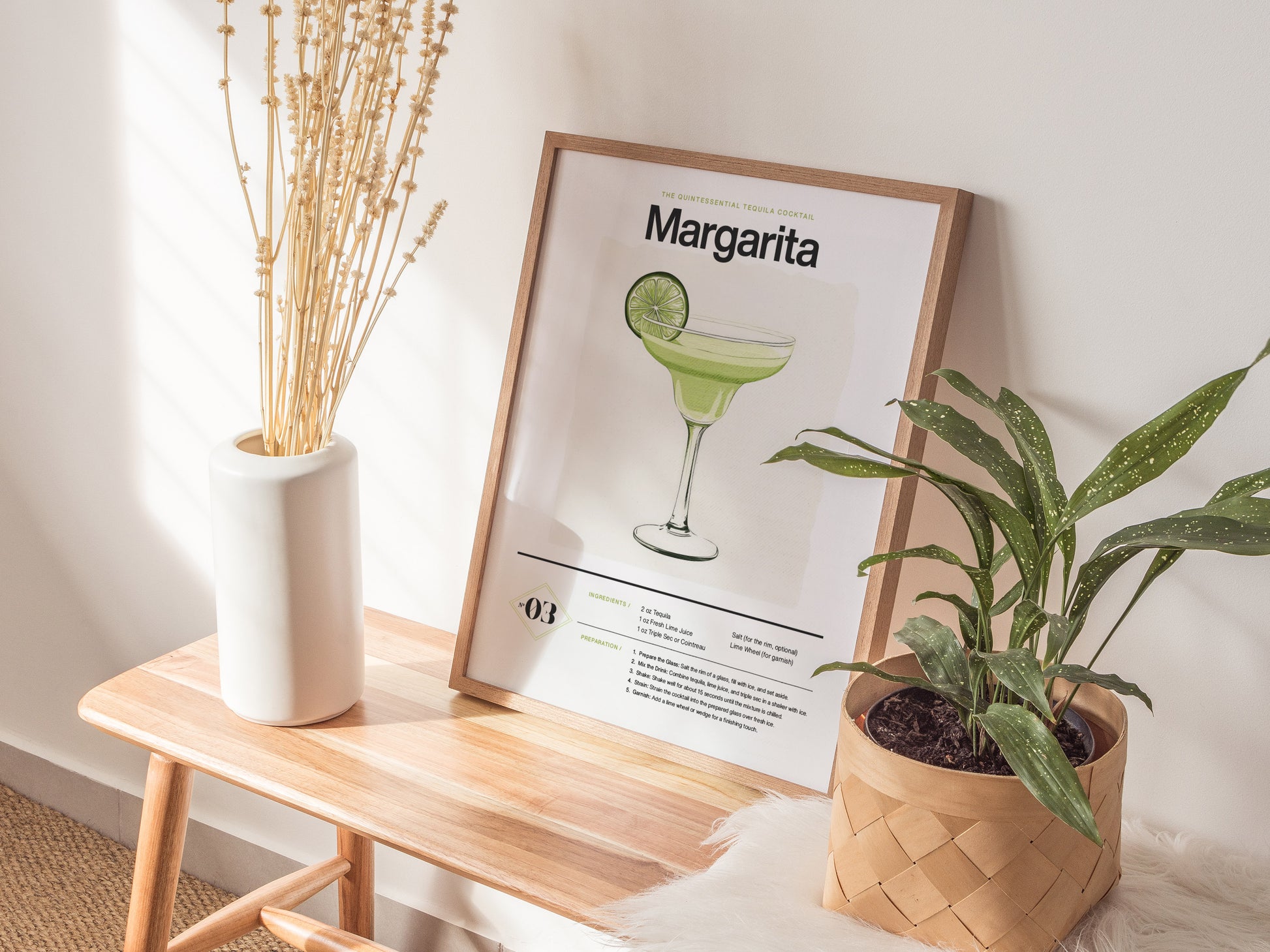 A framed margarita cocktail poster rests on a wooden bench, flanked by a white vase with dried stems and a potted green plant. Sunlight casts subtle shadows.