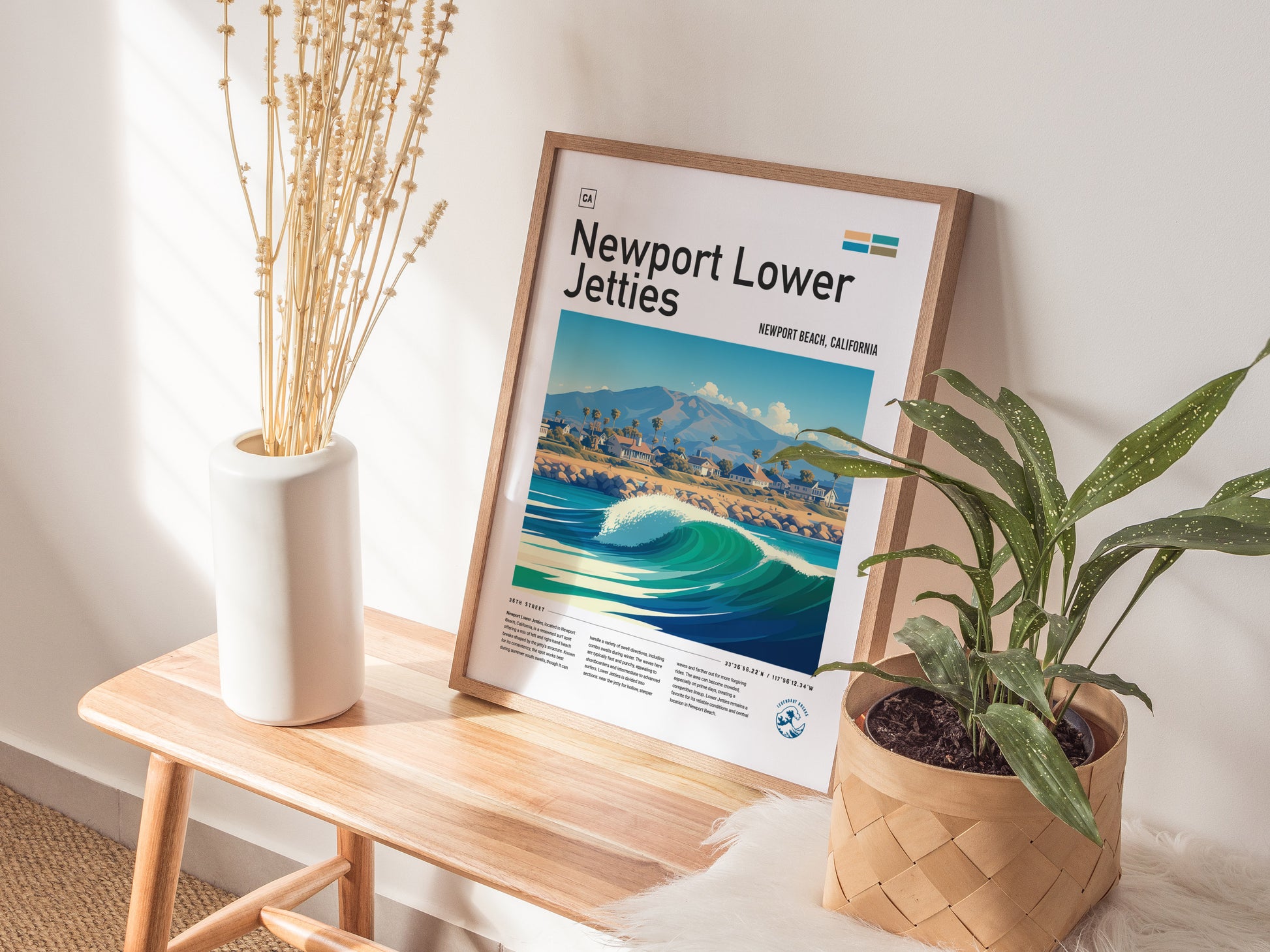 A framed Newport Lower Jetties poster rests on a wooden bench, accompanied by a white vase with dried flowers and a potted green plant, in a sunlit room.