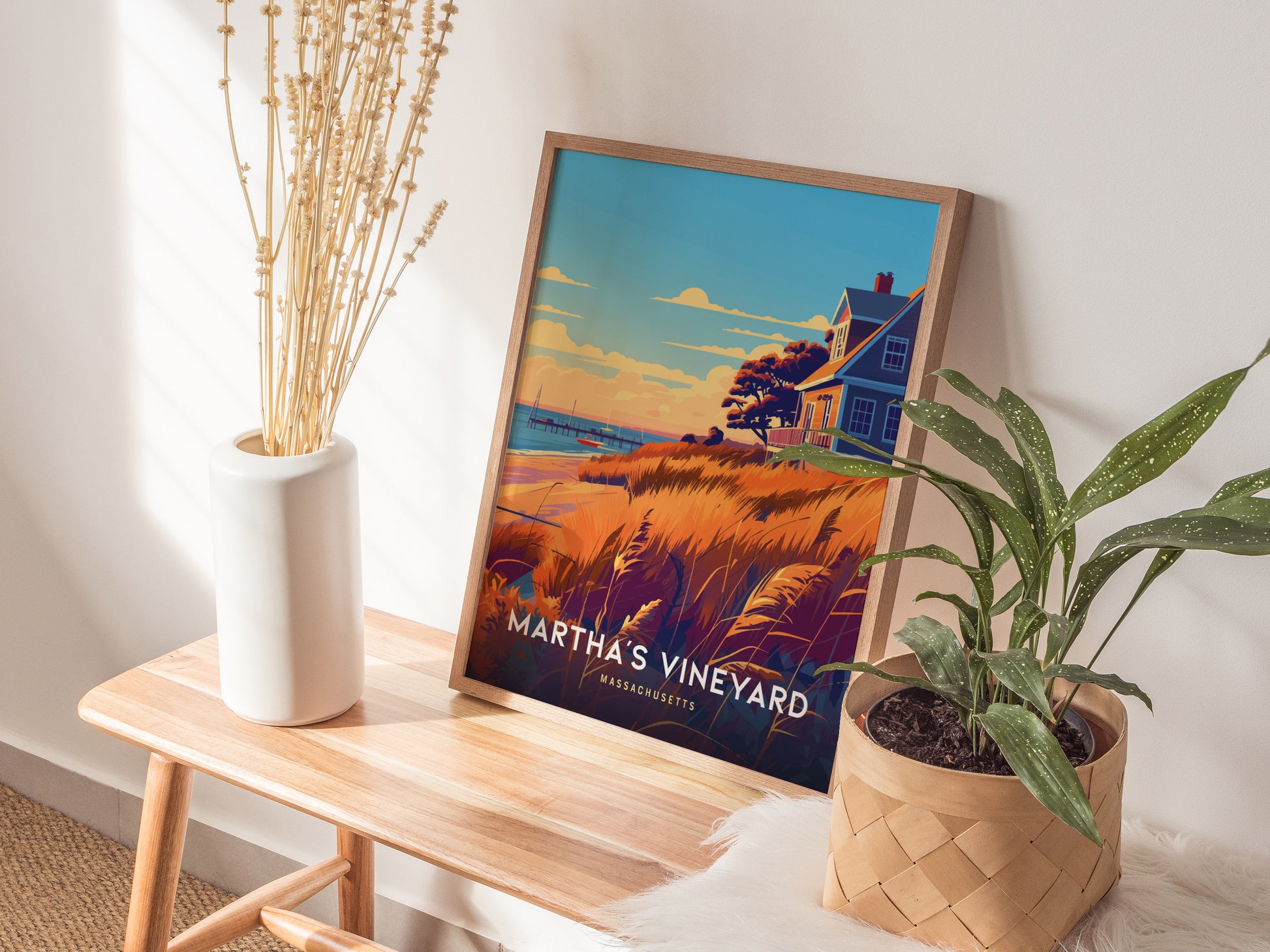 A framed Martha’s Vineyard poster rests on a wooden bench beside a white vase with dried flowers and a potted plant, creating a cozy and inviting interior setting.