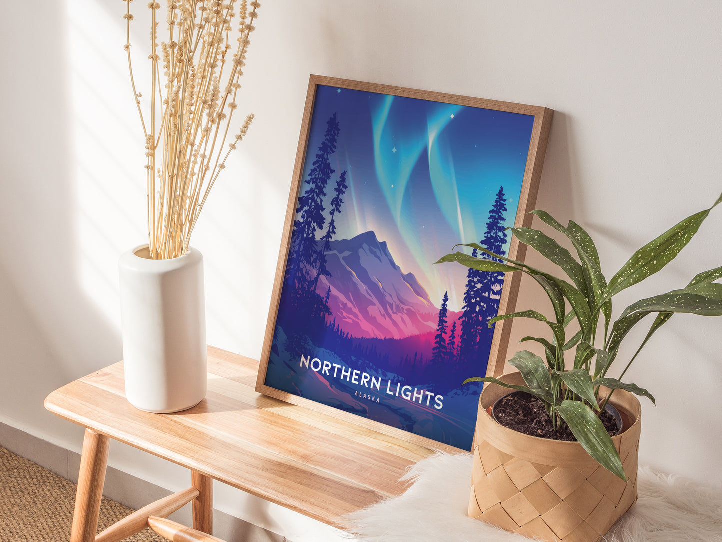 A framed Northern Lights Alaska poster rests on a wooden bench, accompanied by a white vase with dried flowers and a potted green plant, in a softly lit room.