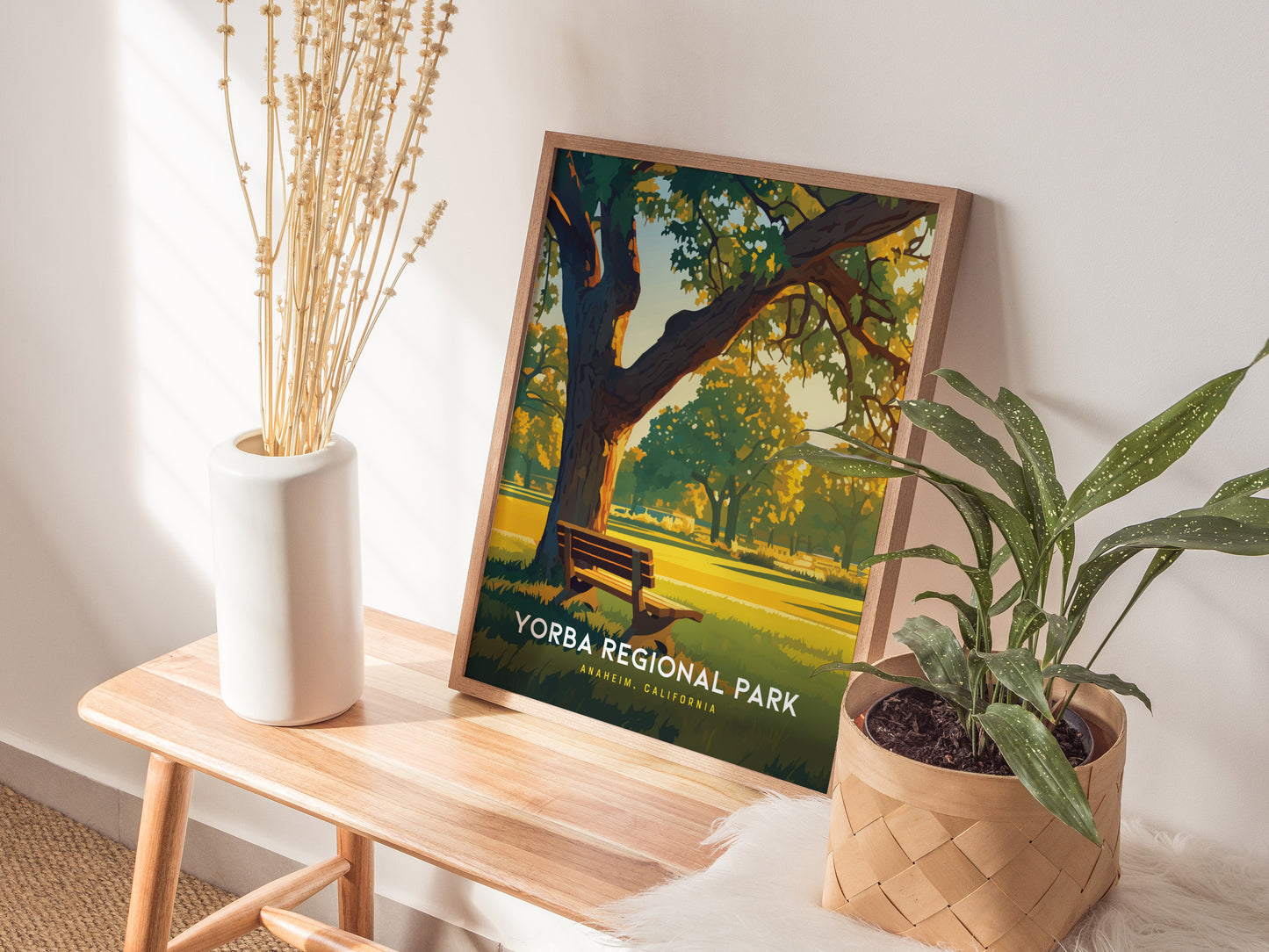 A Yorba Regional Park poster rests on a wooden bench, depicting a serene park scene with trees and a bench. It is surrounded by a white vase with dried flowers.