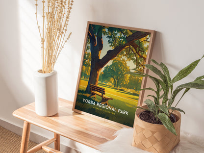 A Yorba Regional Park poster rests on a wooden bench, depicting a serene park scene with trees and a bench. It is surrounded by a white vase with dried flowers.