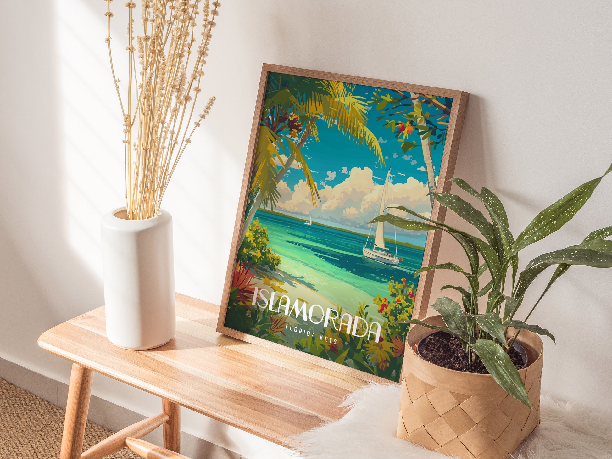 A framed Islamorada poster featuring a vibrant tropical scene with turquoise waters and sailboats is displayed on a wooden bench, accompanied by a vase of dried flowers and a potted.