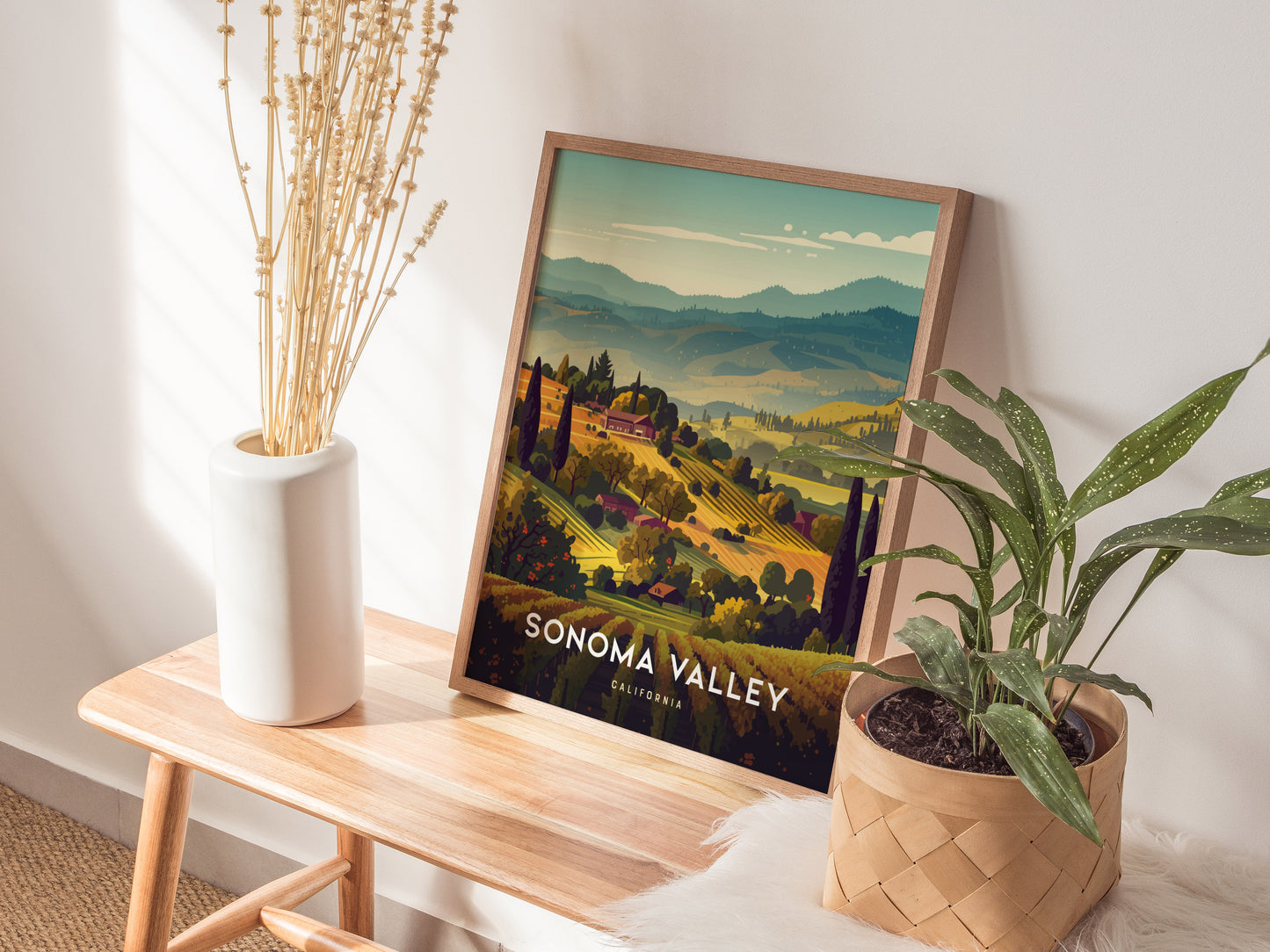 A framed Sonoma Valley poster showcasing vineyard hills is placed on a wooden bench, accompanied by a white vase with dried flowers and a potted plant.
