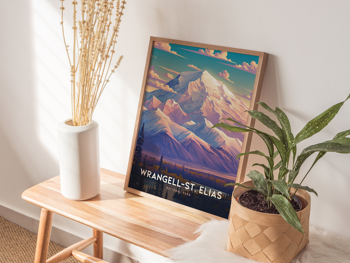 A framed Wrangell-St. Elias National Park poster featuring colorful mountain art rests on a wooden bench beside a white vase with dried flowers and a potted green plant.