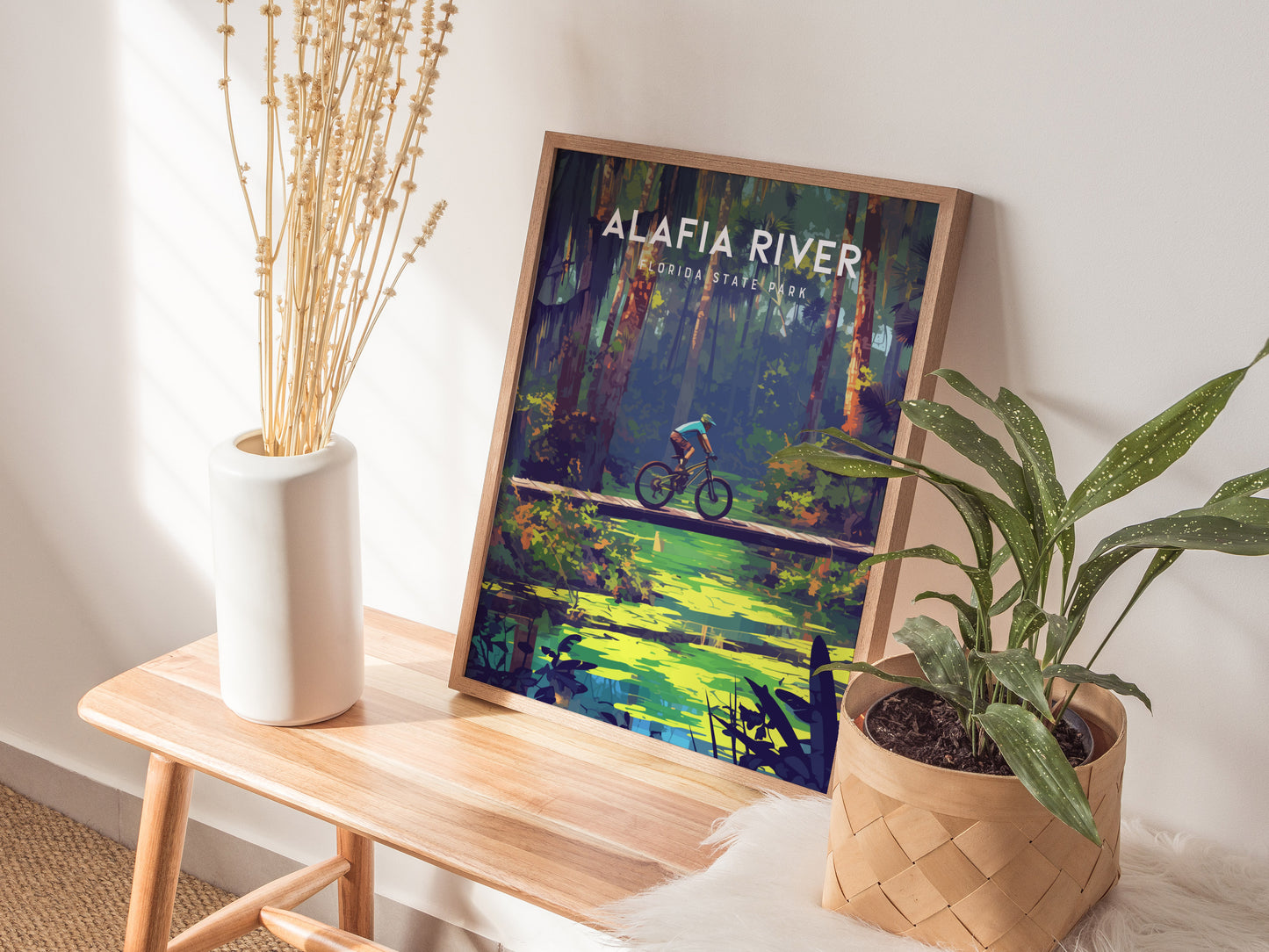 A framed Alafia River State Park poster depicting a mountain biker on a trail is displayed on a wooden bench, accompanied by a white vase with dried flowers and a.