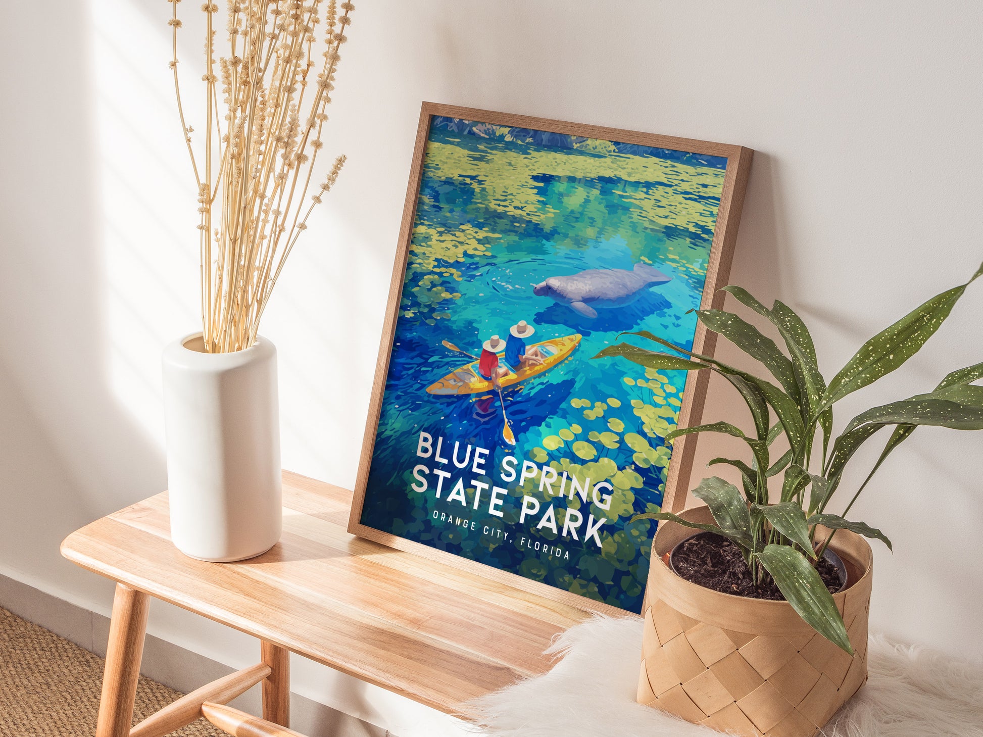 A framed poster of Blue Spring State Park, featuring kayakers and a manatee, rests on a wooden bench beside a white vase with dried flowers and a potted plant.