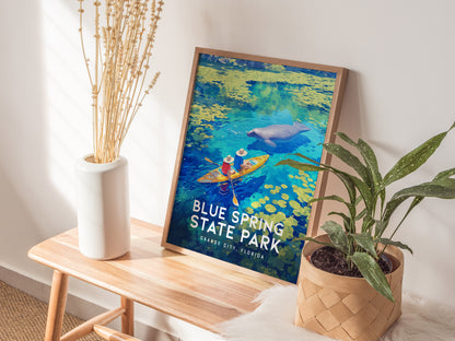 A framed poster of Blue Spring State Park, featuring kayakers and a manatee, rests on a wooden bench beside a white vase with dried flowers and a potted plant.