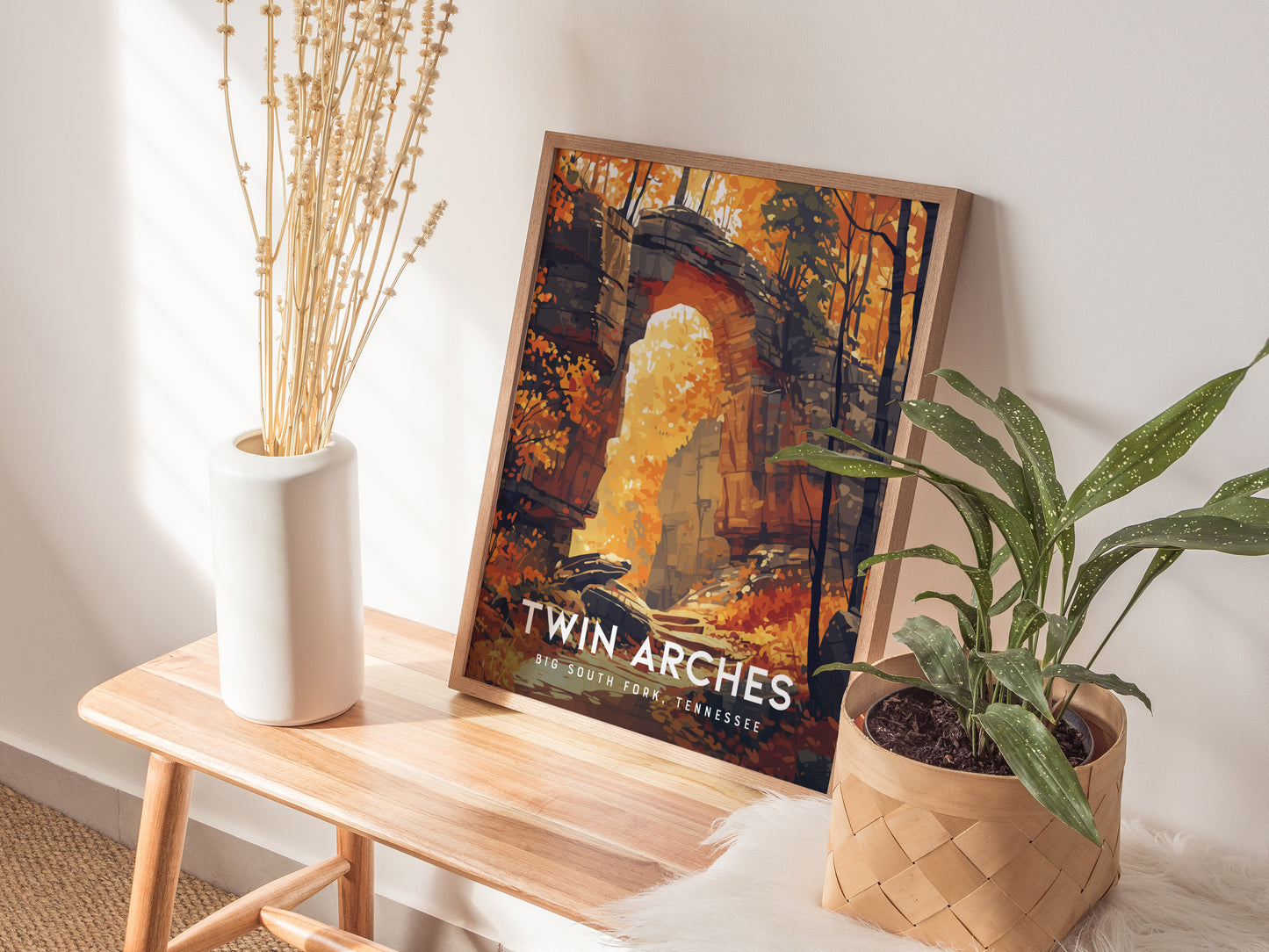 A framed Twin Arches poster rests on a wooden bench beside a white vase and a potted plant, illuminated by natural light.